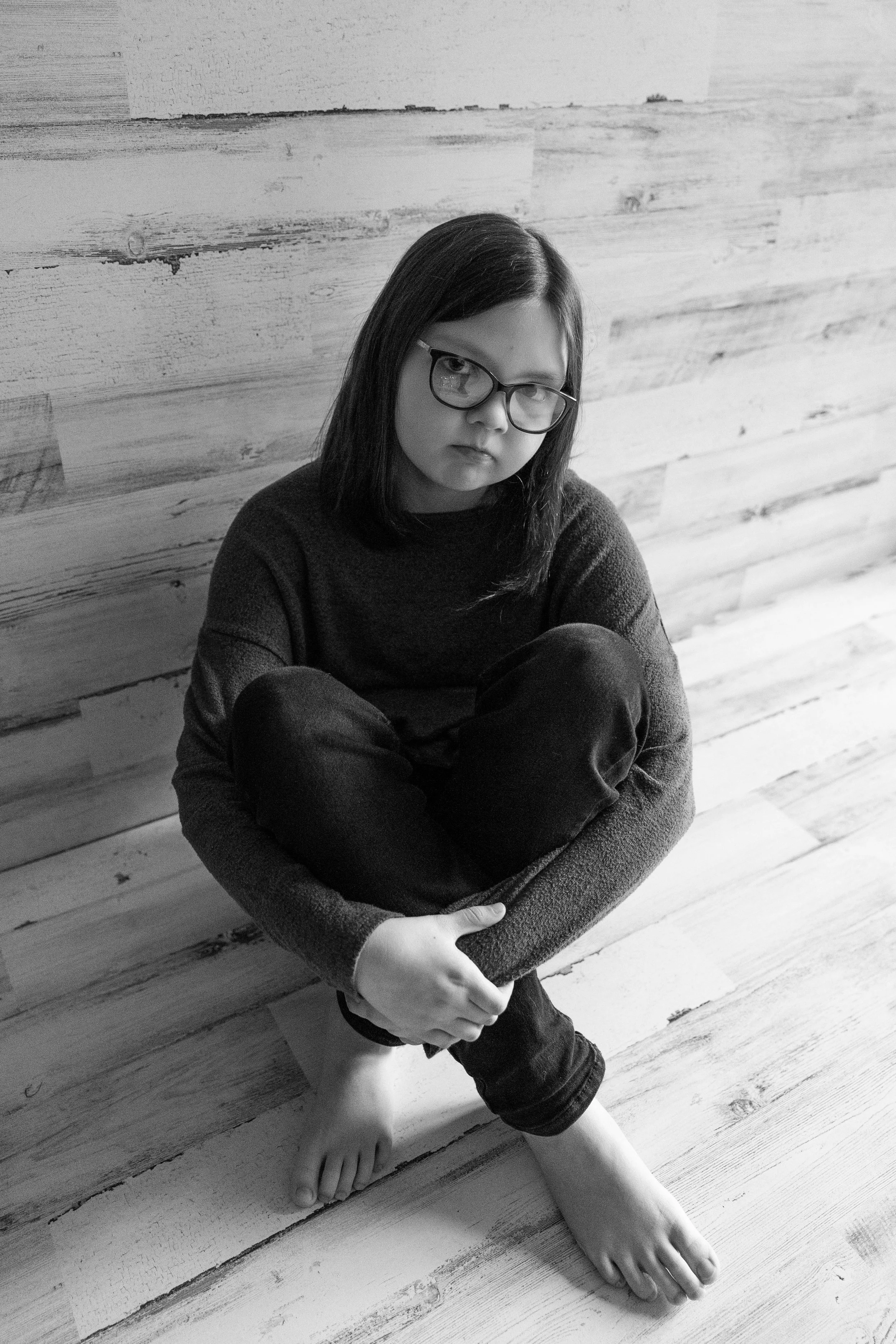 A young girl with glasses sitting on a wooden floor, with her arms wrapped around her knees, looking at the camera.