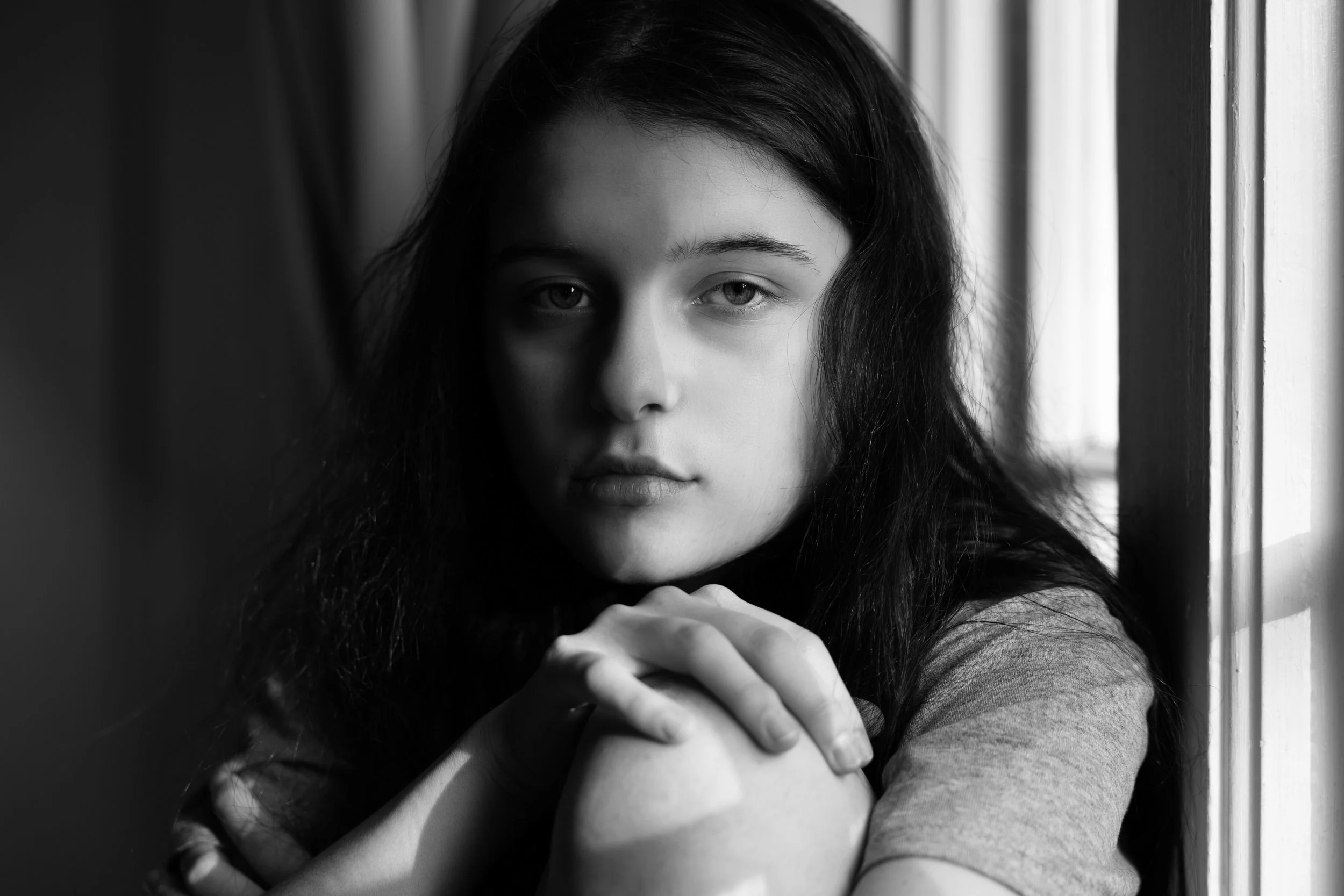 Black and white portrait of a young woman with long dark hair, leaning on her arms near a window, looking directly at the camera.