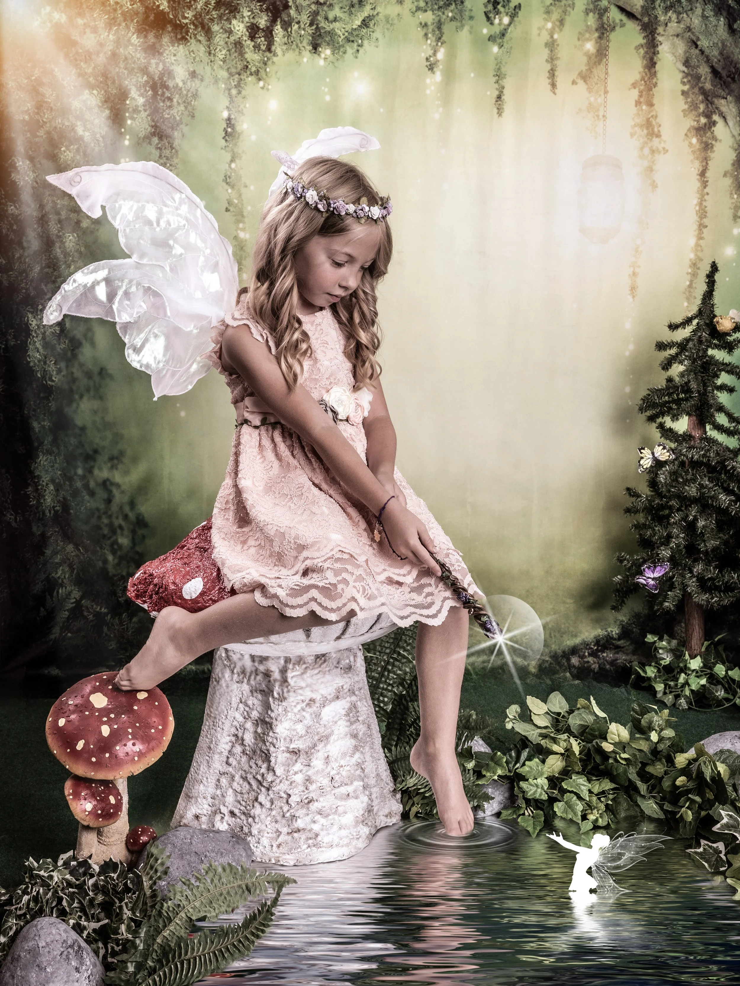 A young girl dressed as a fairy with wings and a floral headband, sitting on a tree stump trotting her foot in a pond, with a fairy figurine floating on the water, surrounded by lush greenery and mushrooms.