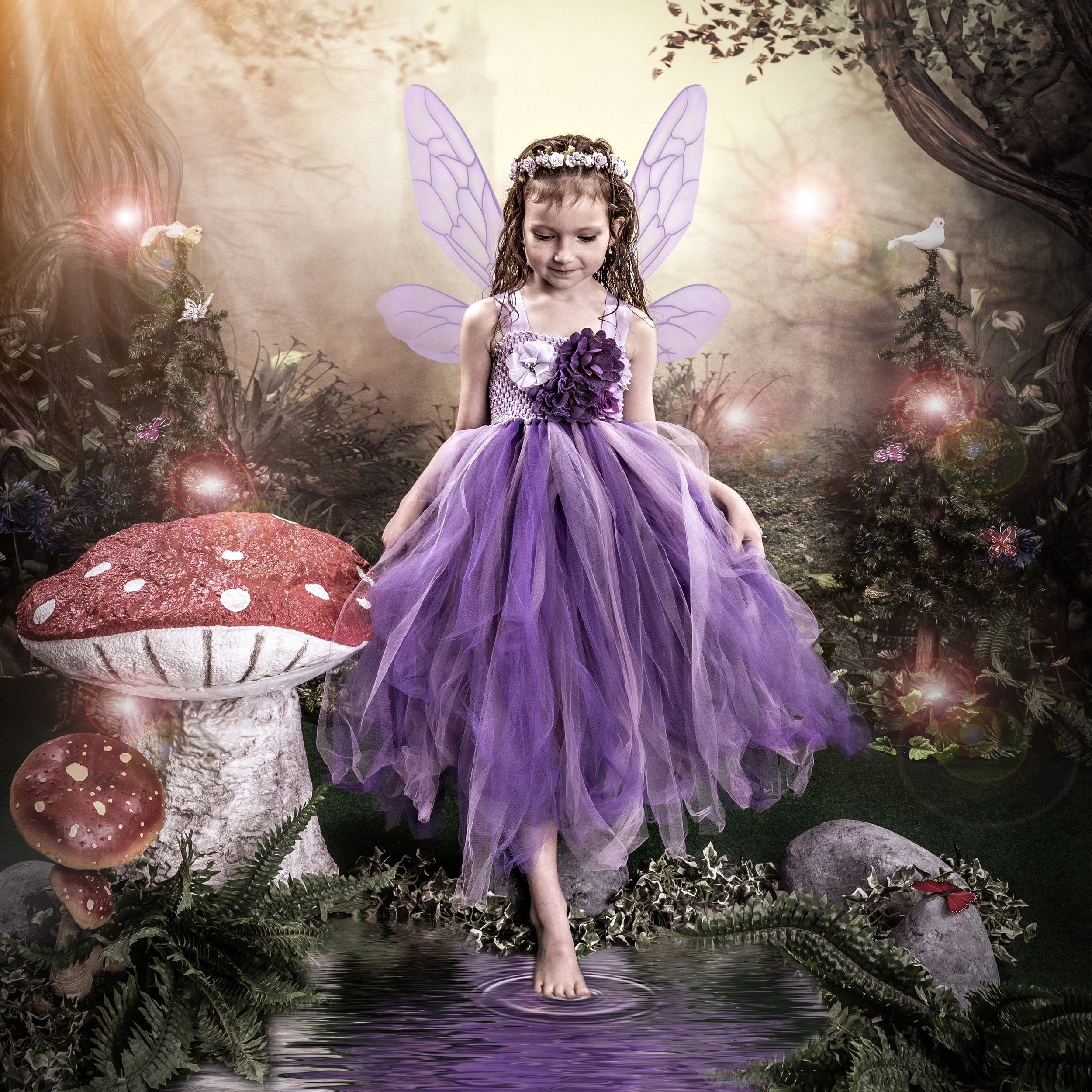 Young girl dressed as a fairy with wings, standing beside a large red mushroom with white spots in a magical forest setting.