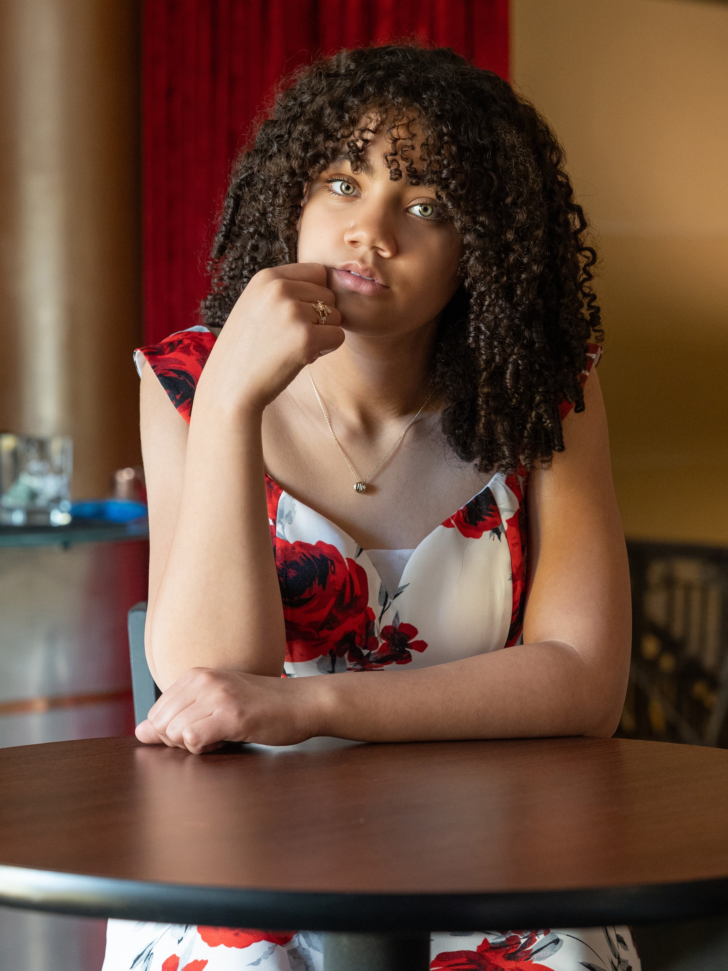 A young woman with curly hair and light skin, wearing a floral dress, sitting at a table with her chin resting on her hand, looking at the camera.