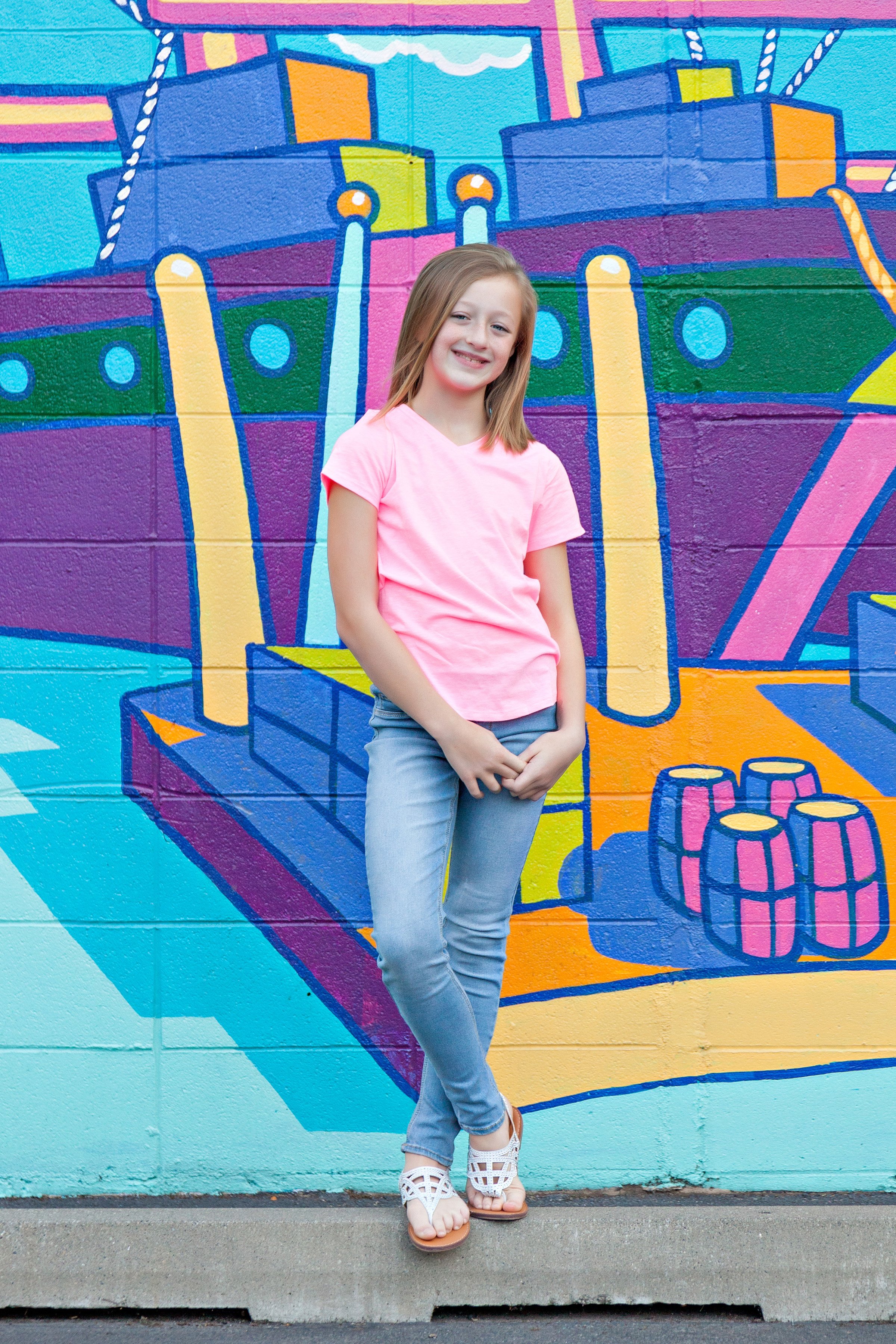 A young girl with blonde hair smiling, wearing a pink t-shirt, gray pants, and white sandals, standing in front of a colorful mural of a boat on a wall.