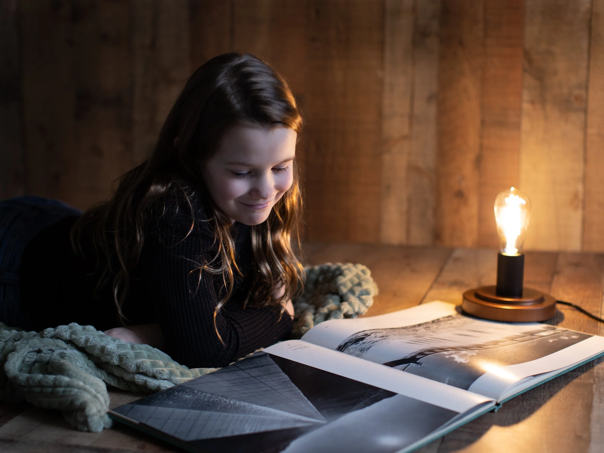 A young girl with long brown hair lying on her stomach, reading a large photo book on a wooden surface. There is a glowing Edison-style bulb lamp on her right, illuminating the scene, with a plush blanket nearby. The background features a wooden wall