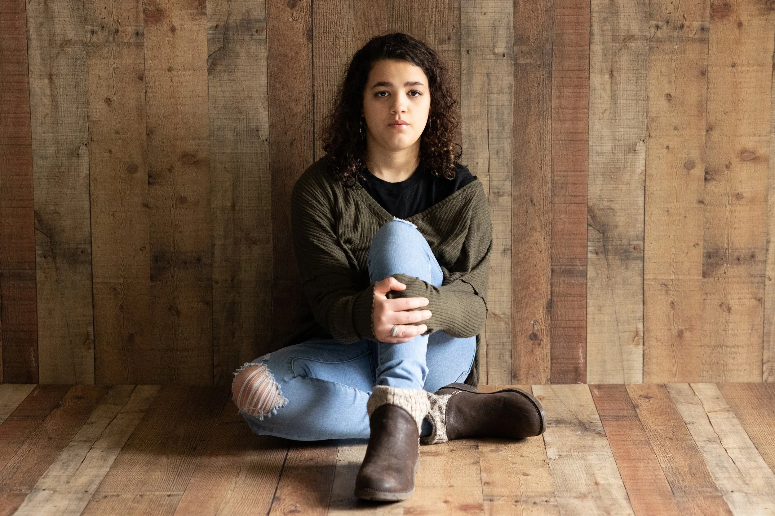 A young woman with curly hair, wearing a green sweater, black shirt, ripped jeans, and brown boots, sitting on a wooden floor against a wooden wall with her knees drawn up, looking directly at the camera.