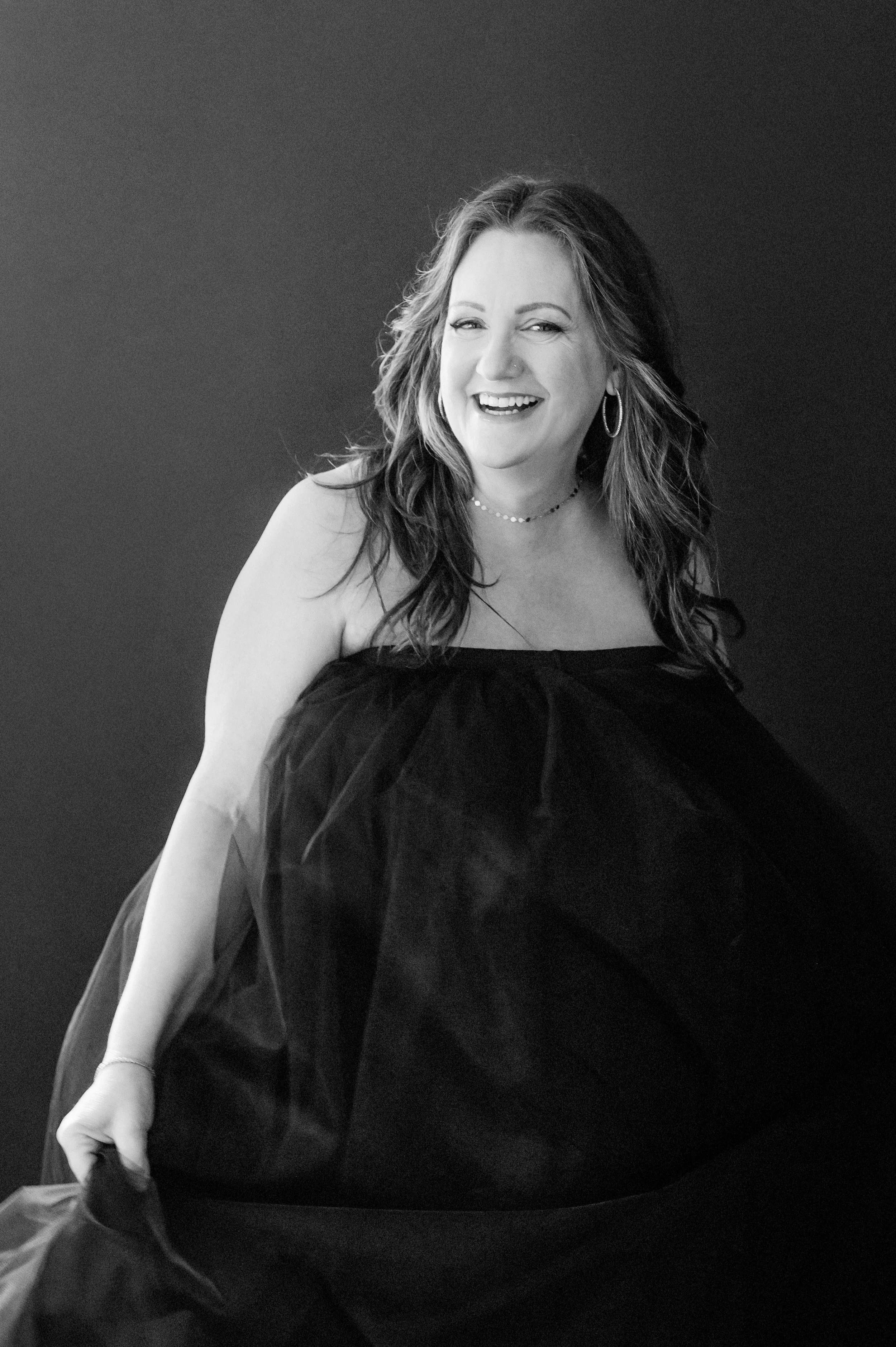 A woman with wavy hair wearing a strapless dress, smiling and looking at the camera, against a dark background.