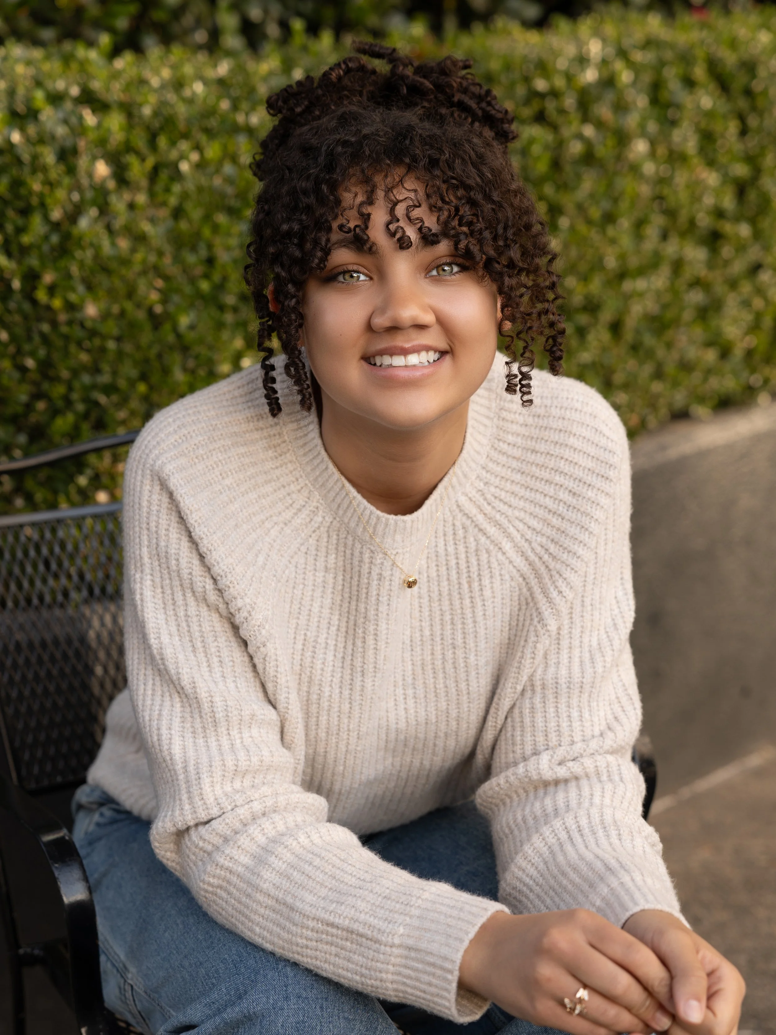 A young woman with curly hair and green eyes smiling while sitting outdoors on a bench, wearing a beige sweater and blue jeans, with greenery in the background.