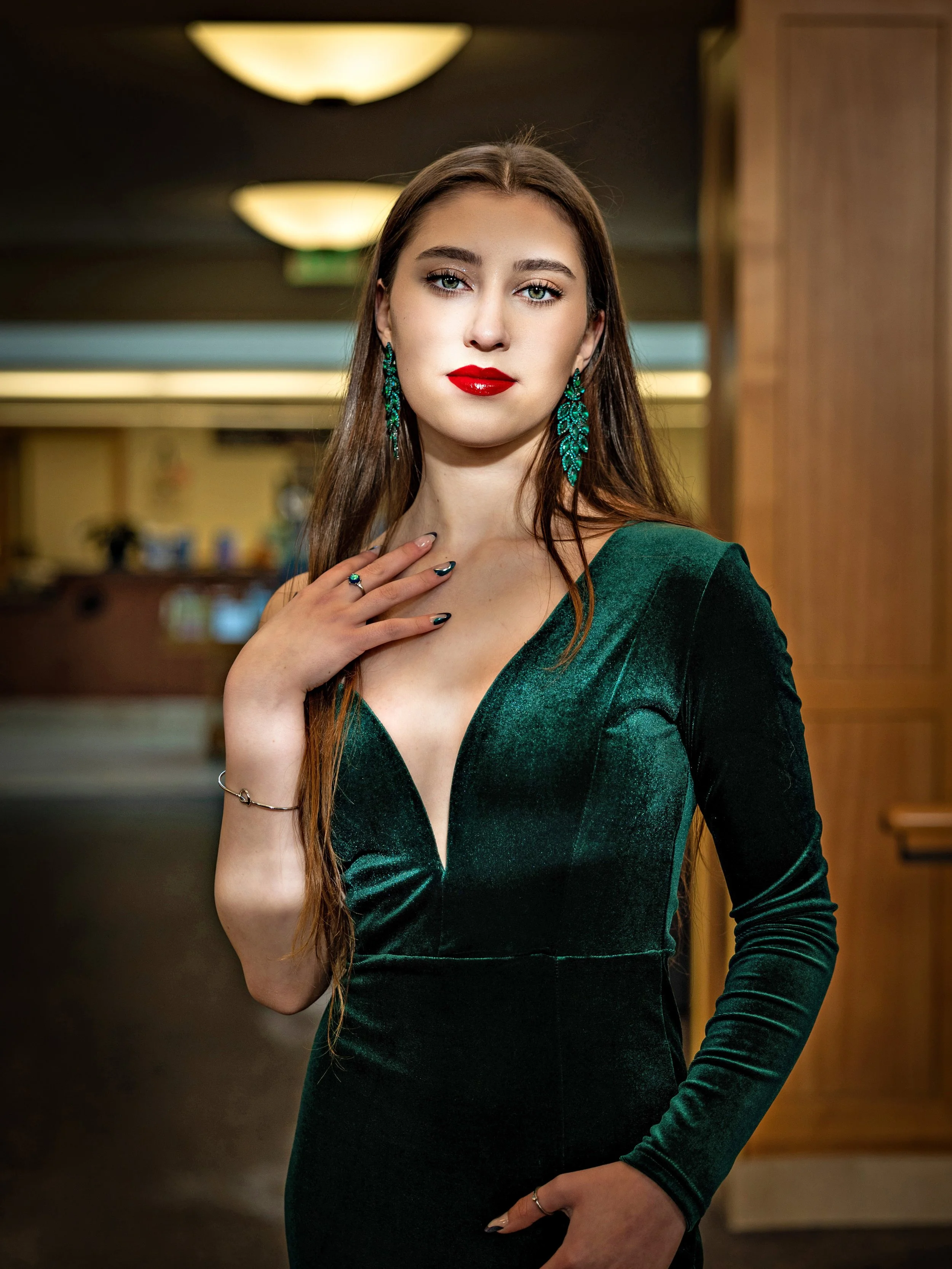 A woman in a dark green velvet dress, with red lipstick and green earrings, standing indoors with a confident expression.