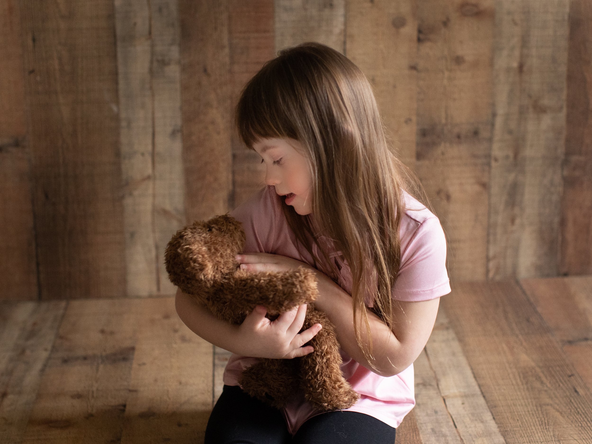 A young girl with long brown hair, wearing a pink shirt, is hugging a brown teddy bear. She is looking down at the teddy bear with a gentle expression, standing against a wooden wall background.