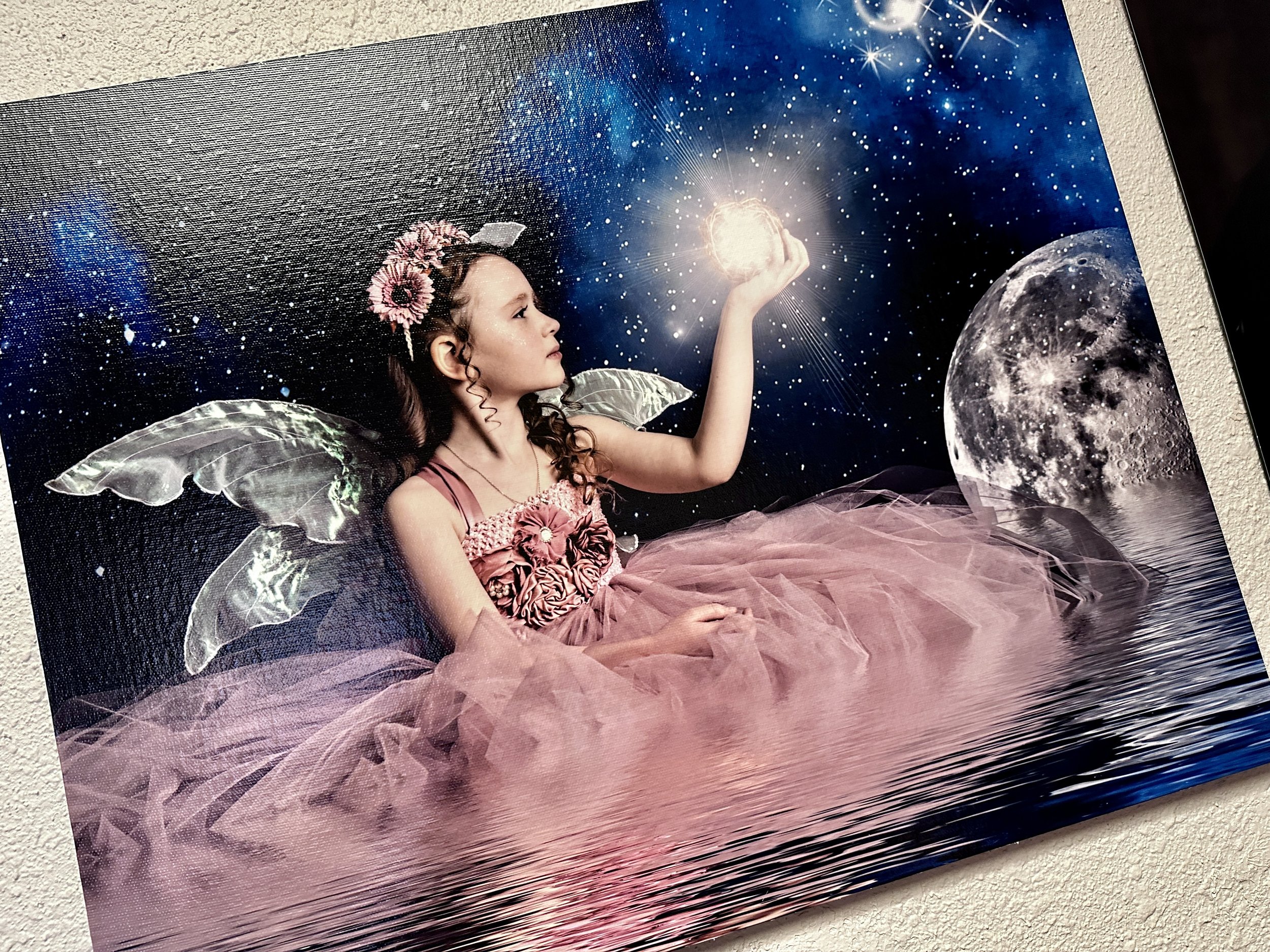 A young girl dressed as a fairy with wings and a pink dress, holding a glowing orb against a starry night background with a full moon and reflection on water.