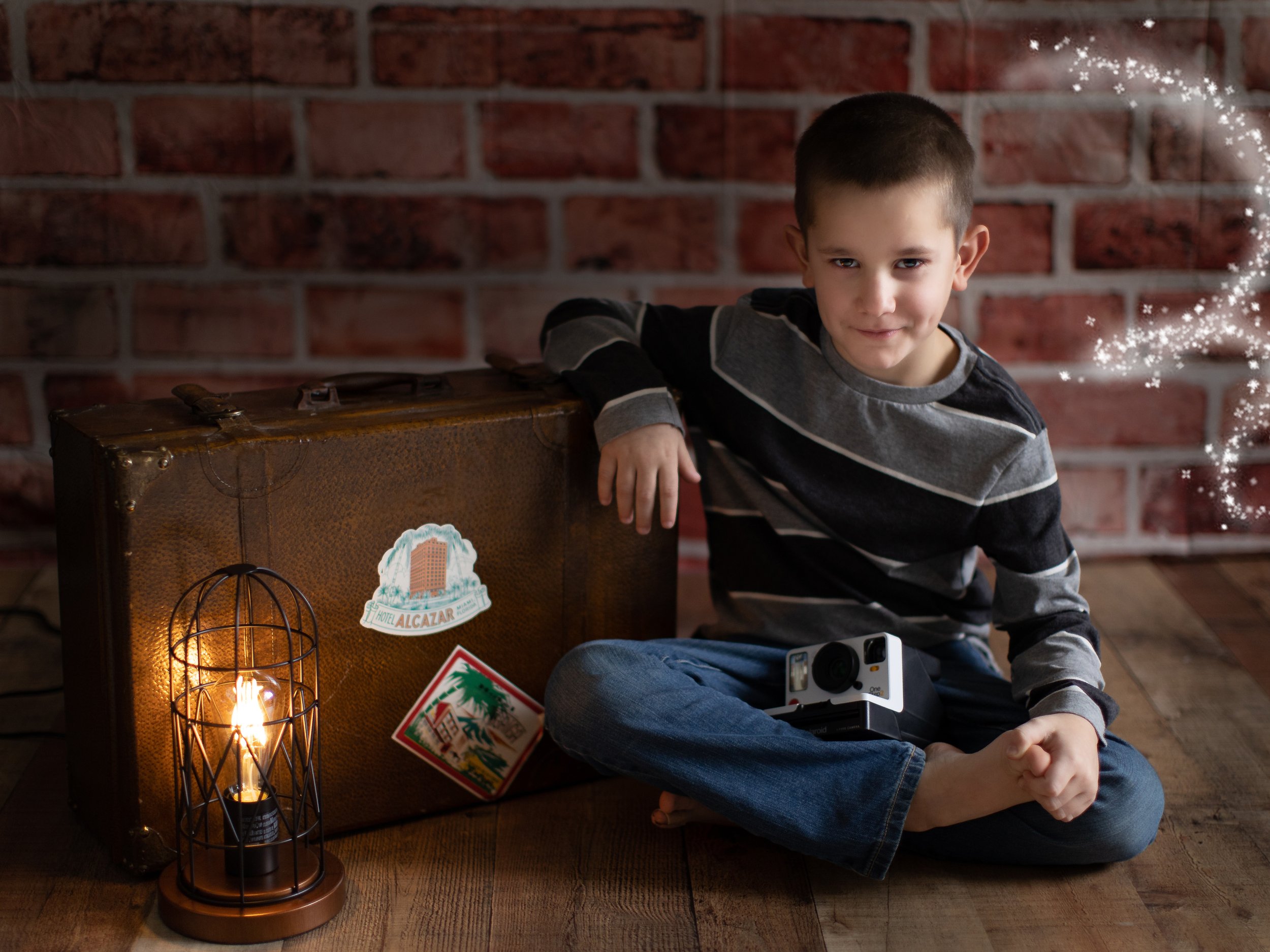 A young boy with short dark hair and a striped long sleeve shirt is sitting on the floor in front of a brick wall, smiling slightly and holding his hands clasped in his lap. In front of him is a vintage-style suitcase with a sticker of a hotel logo a