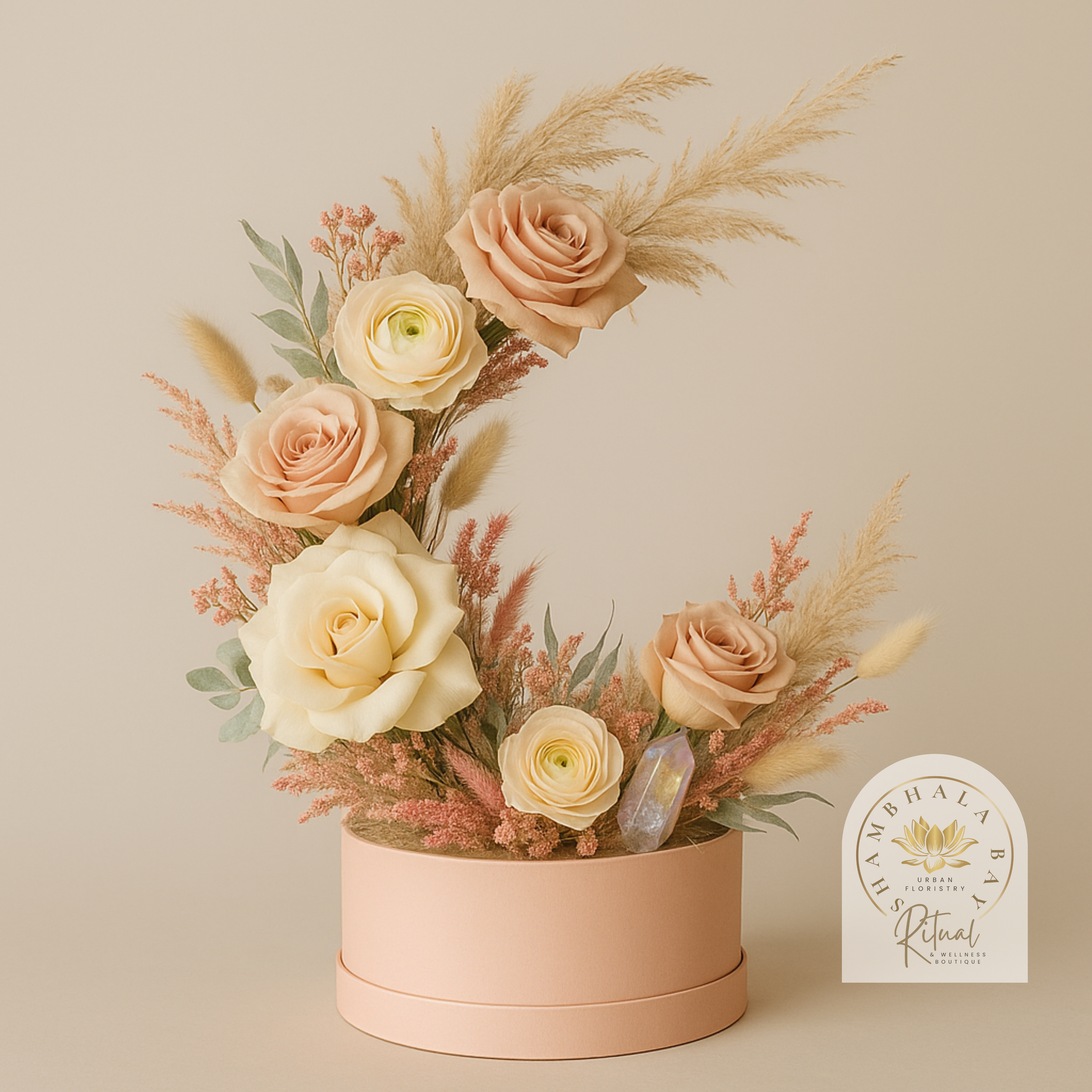 A floral arrangement with roses and dried pampas grass in a pink round hat box. The arrangement includes cream and blush-colored roses, dried pink and beige foliage, and a crystal, Signature Shambhala Bay Design 