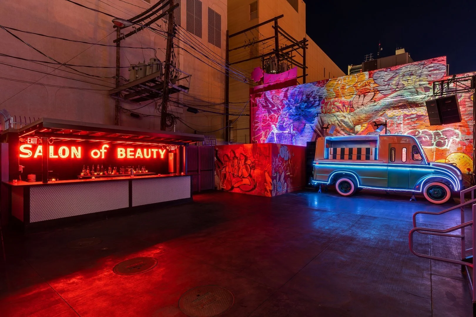 A vibrant urban outdoor scene featuring a bar with a neon sign "Salon of Beauty," graffiti-covered walls, and a retro truck with blue neon lights at night.
