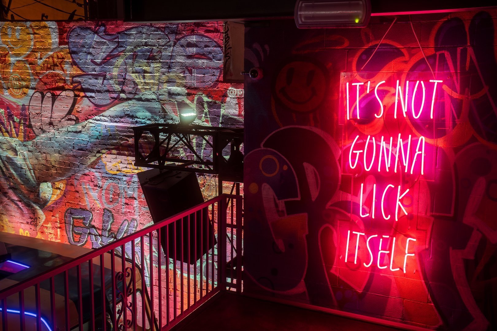 Neon sign with 'IT'S NOT GONNA LICK ITSELF' on a graffiti-covered wall in a dimly lit space.