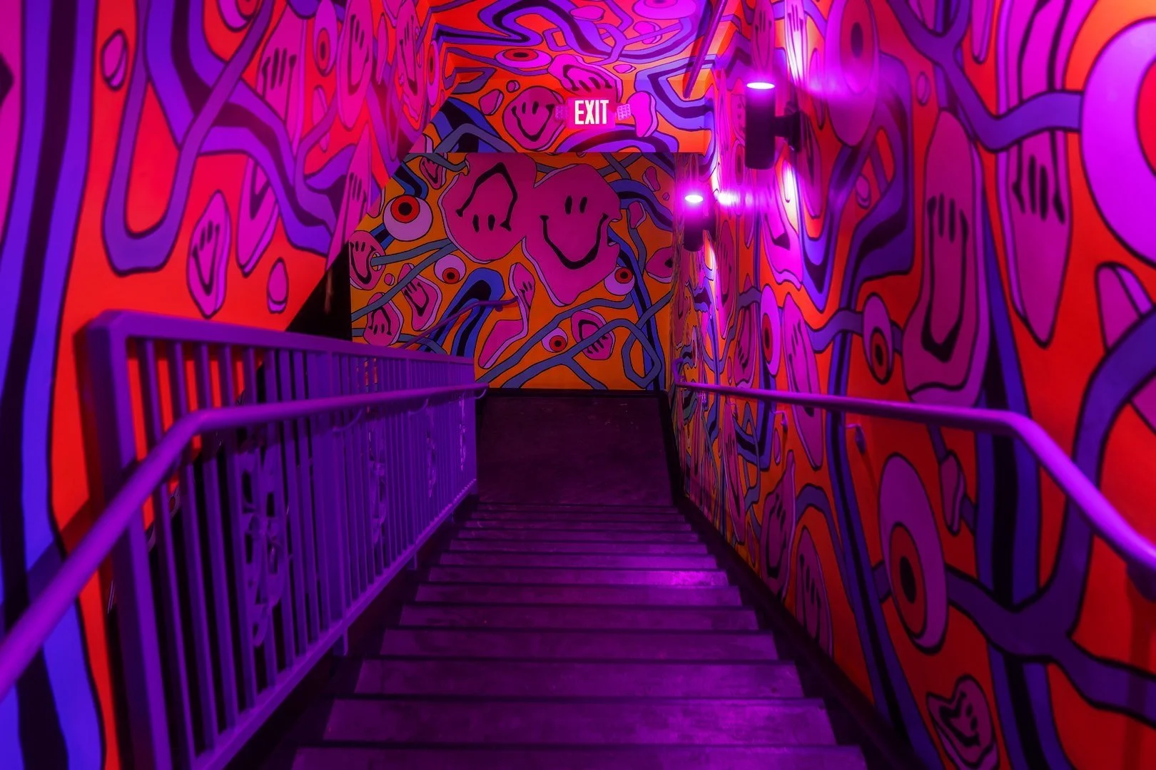 Colorful mural-covered staircase with purple lighting and an exit sign.