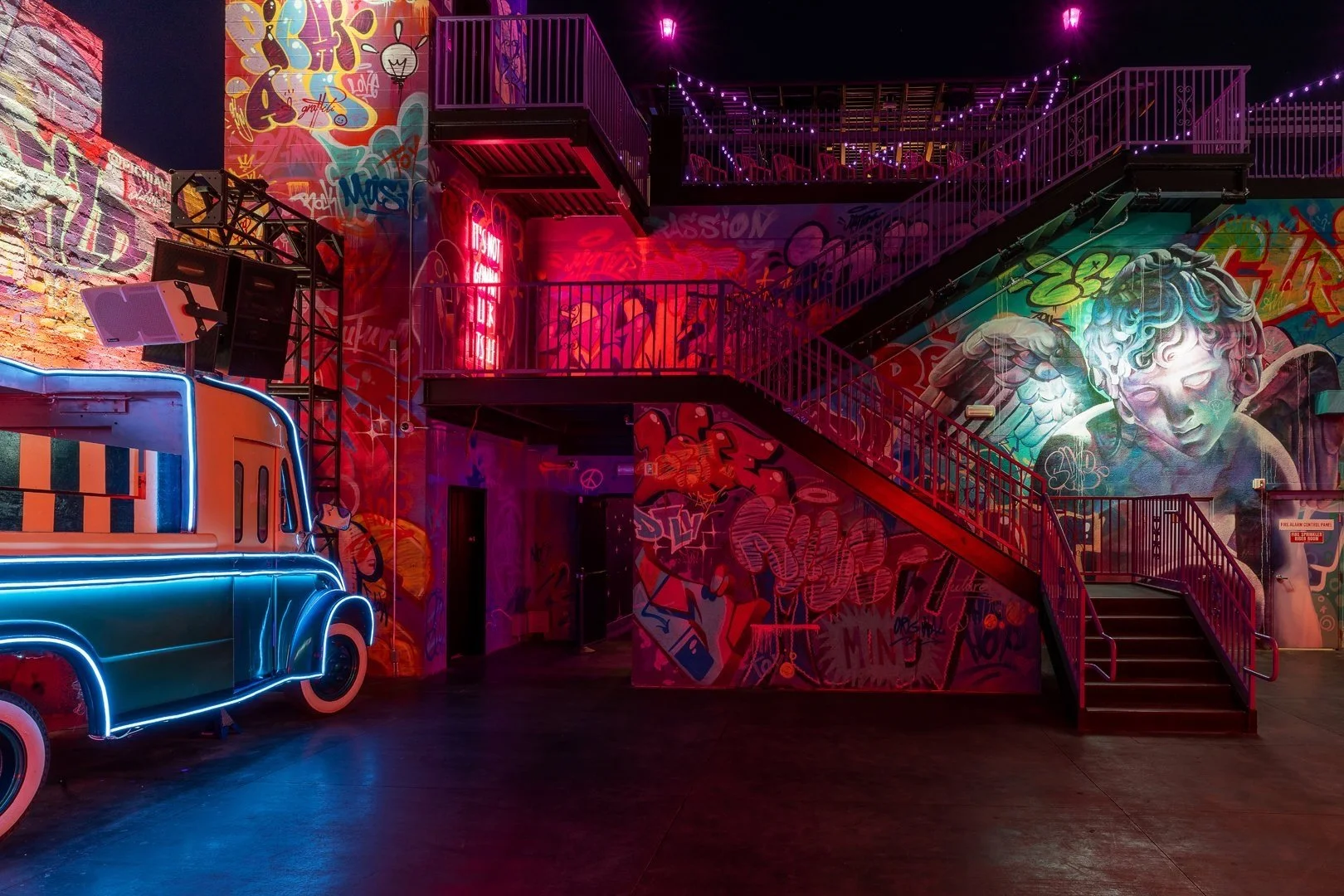 Interior of a colorful urban setting with graffiti-covered walls, neon lighting, and metal stairs.
