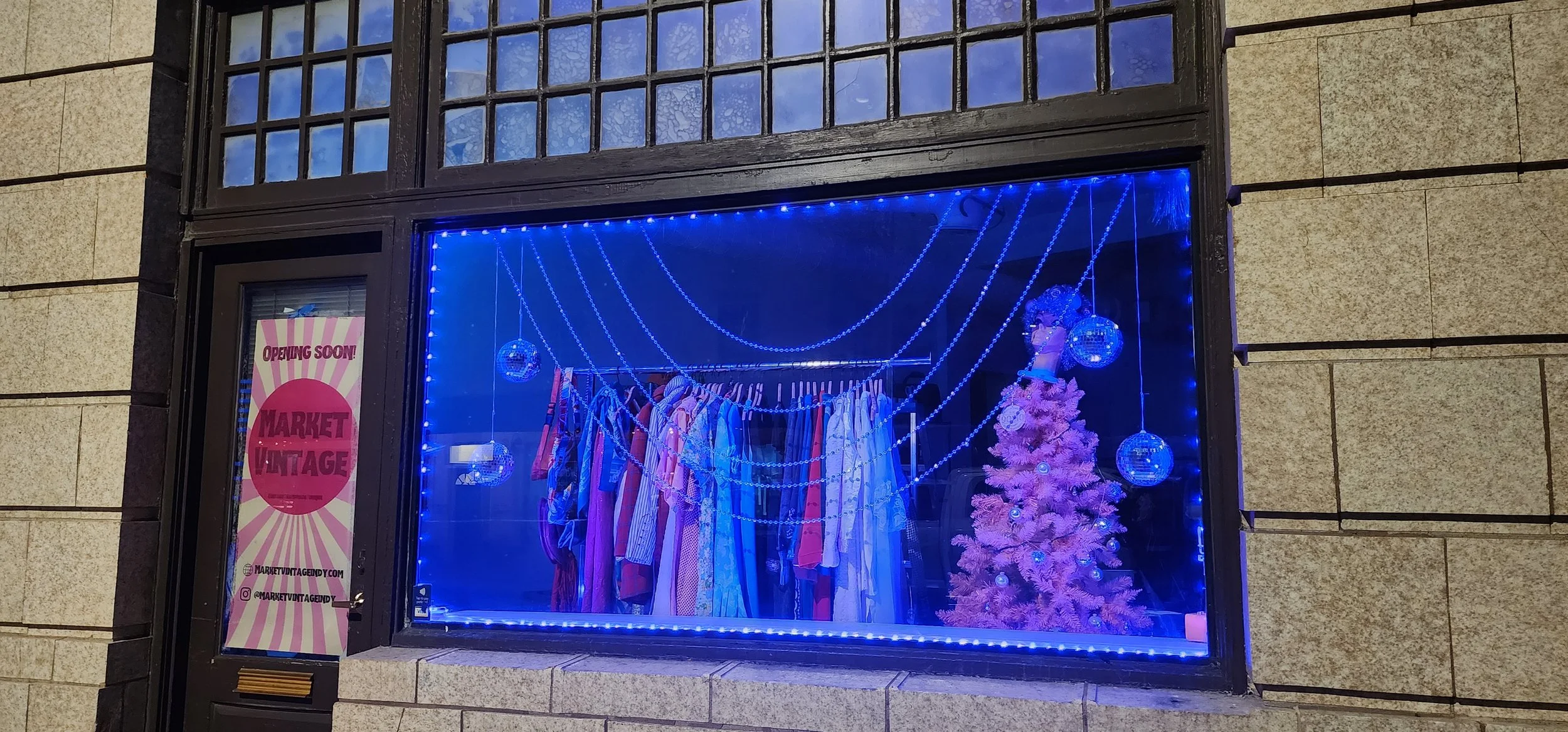 Storefront window display with pink Christmas tree, hanging disco balls, and colorful vintage clothing under blue LED lights. Sign reads "Opening Soon! Market Vintage."