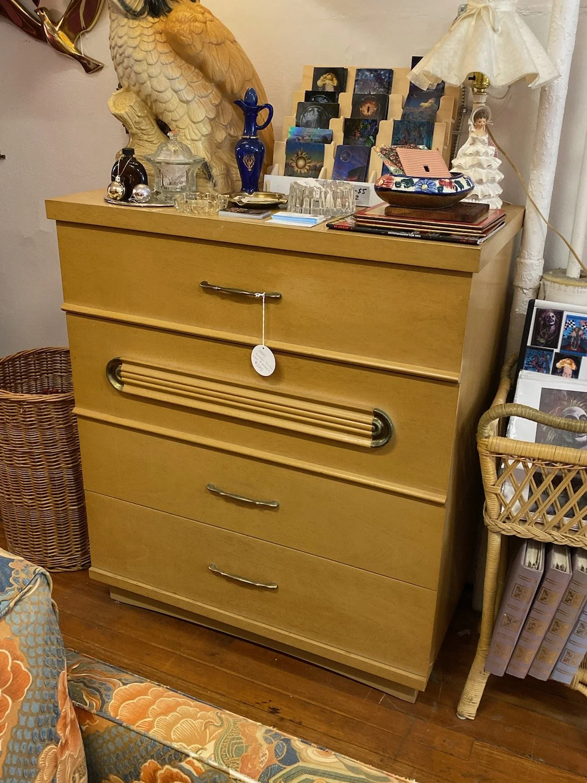 A wooden chest of drawers with decorative items, including a large ceramic bird, blue glass vases, and a lamp with a fabric shade on top. A display of colorful postcards or greeting cards is also present. The chest is situated next to a wicker basket