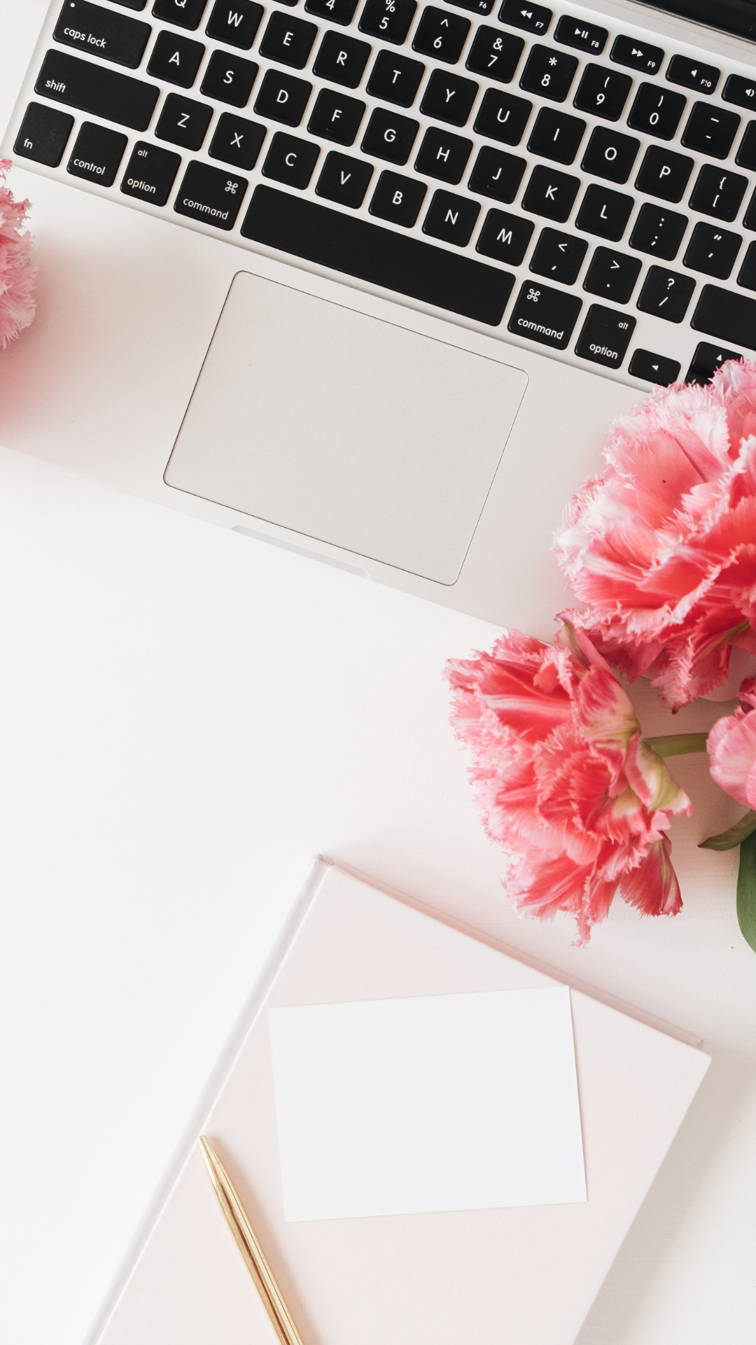 Laptop keyboard with pink tulips, notebook, and gold pen on a white desk.
