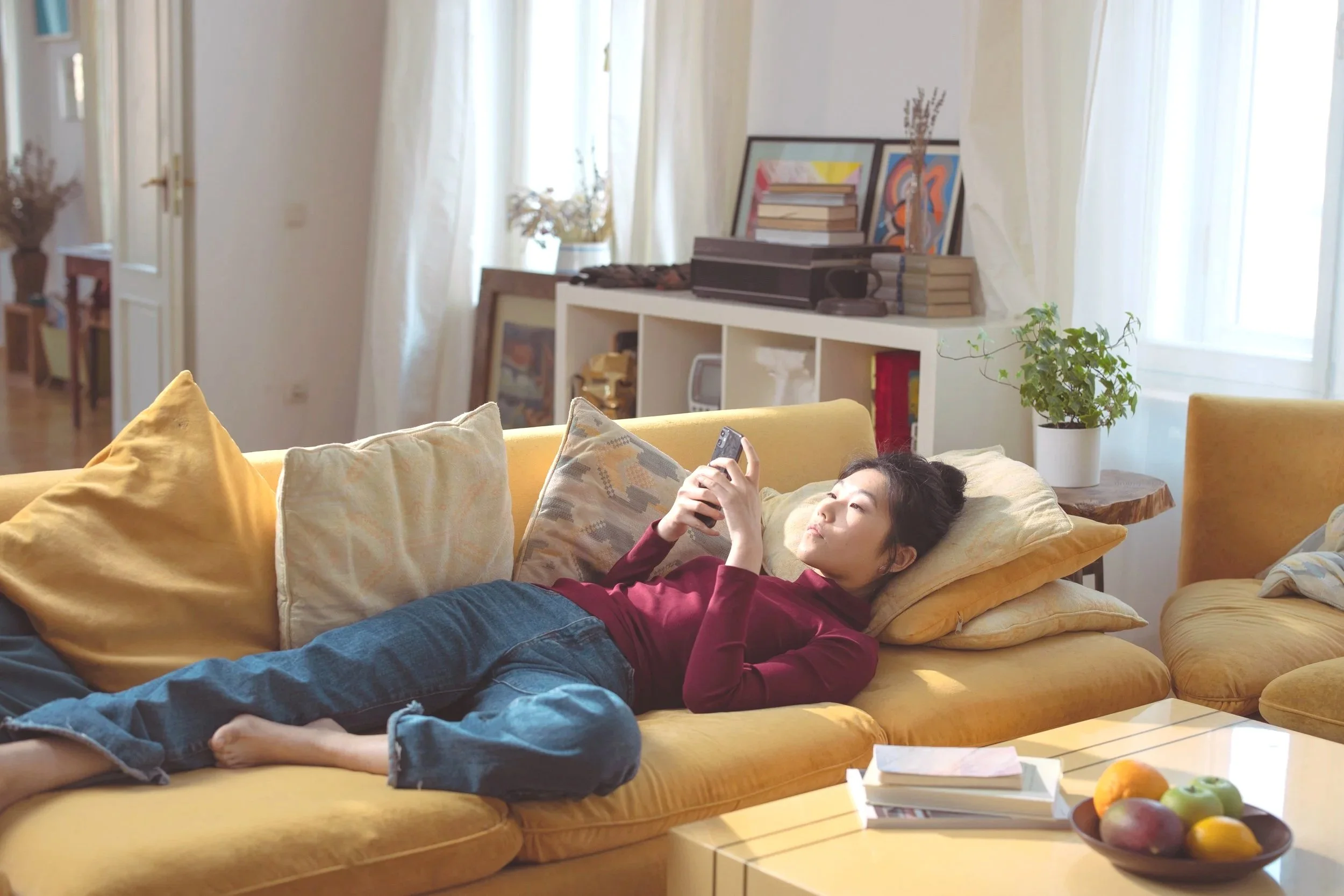 An Asian woman lies down on a couch with her phone, ready to join a virtual therapy session.