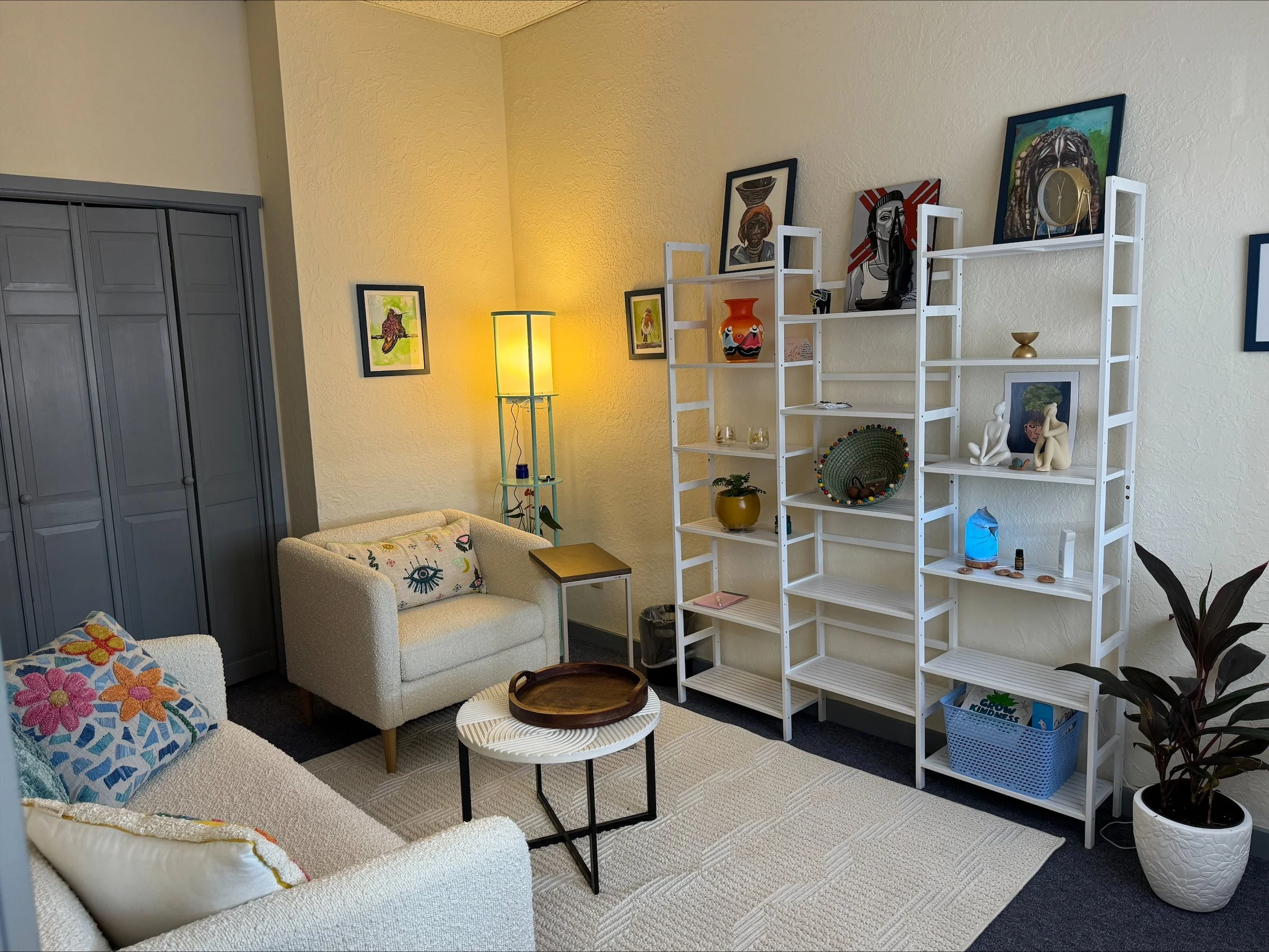 A cozy living room with white sofas, colorful pillows, a modern coffee table, a yellow floor lamp, a tall white bookshelf with decorative items, artwork on the walls, and a large potted plant in a white planter.