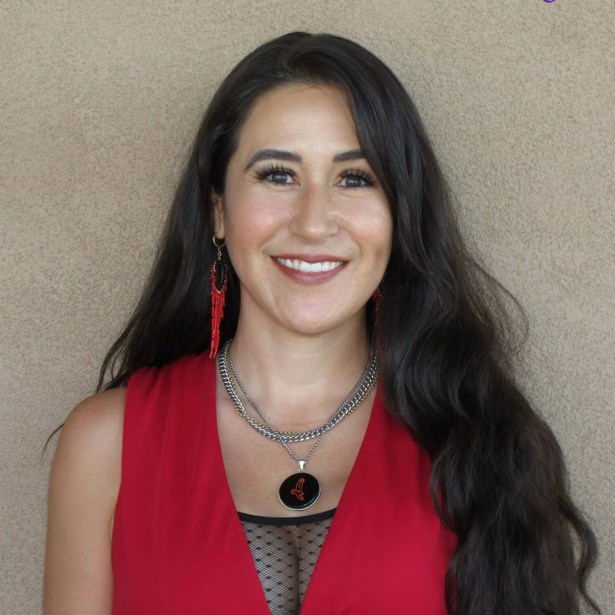 A woman with long, wavy dark hair wearing red beaded earrings and a colorful embroidered top, smiling at the camera.