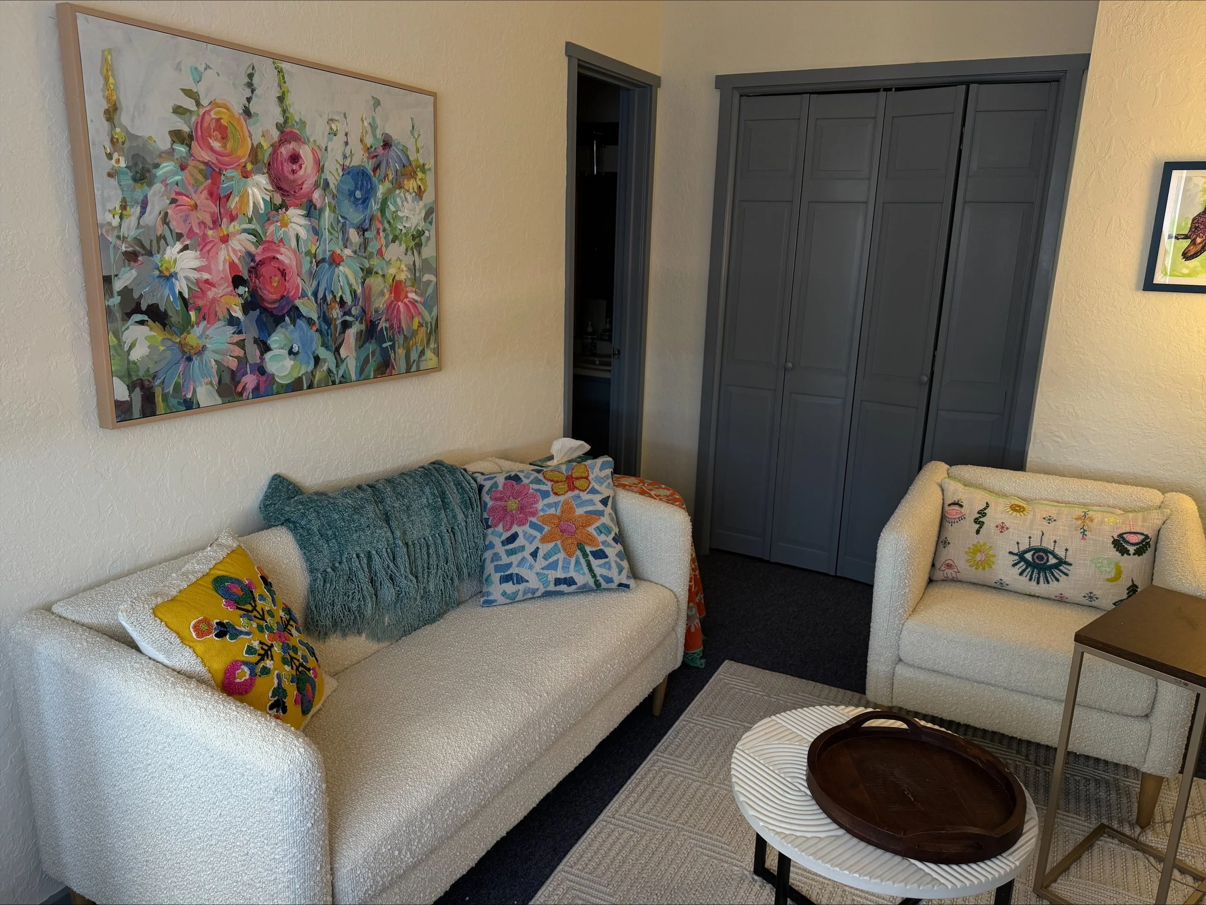 Living room with cream-colored sofa and matching armchair, colorful pillows, large floral painting on wall, dark gray closet doors, side table, and tray on coffee table.
