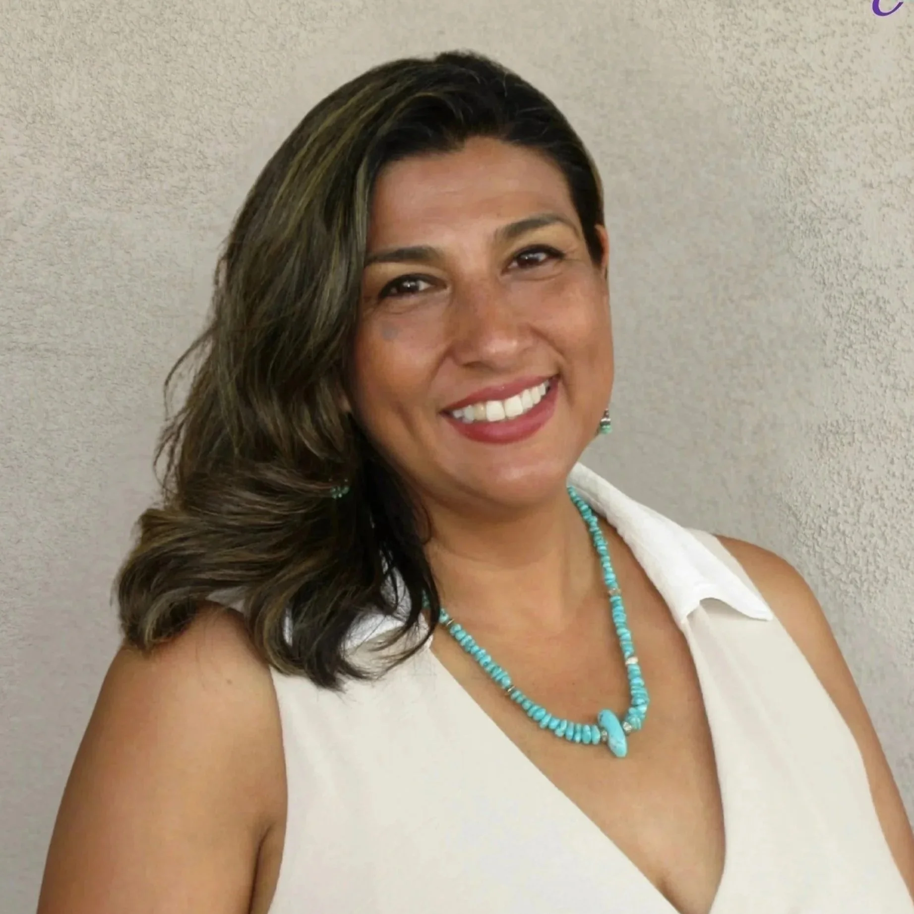 A smiling woman with shoulder-length dark hair, wearing a white sleeveless dress, a turquoise necklace, earrings, and a watch, standing in front of a beige textured wall with a logo in the top right corner that reads 'Grace Integrated Solutions' with a butterfly graphic.