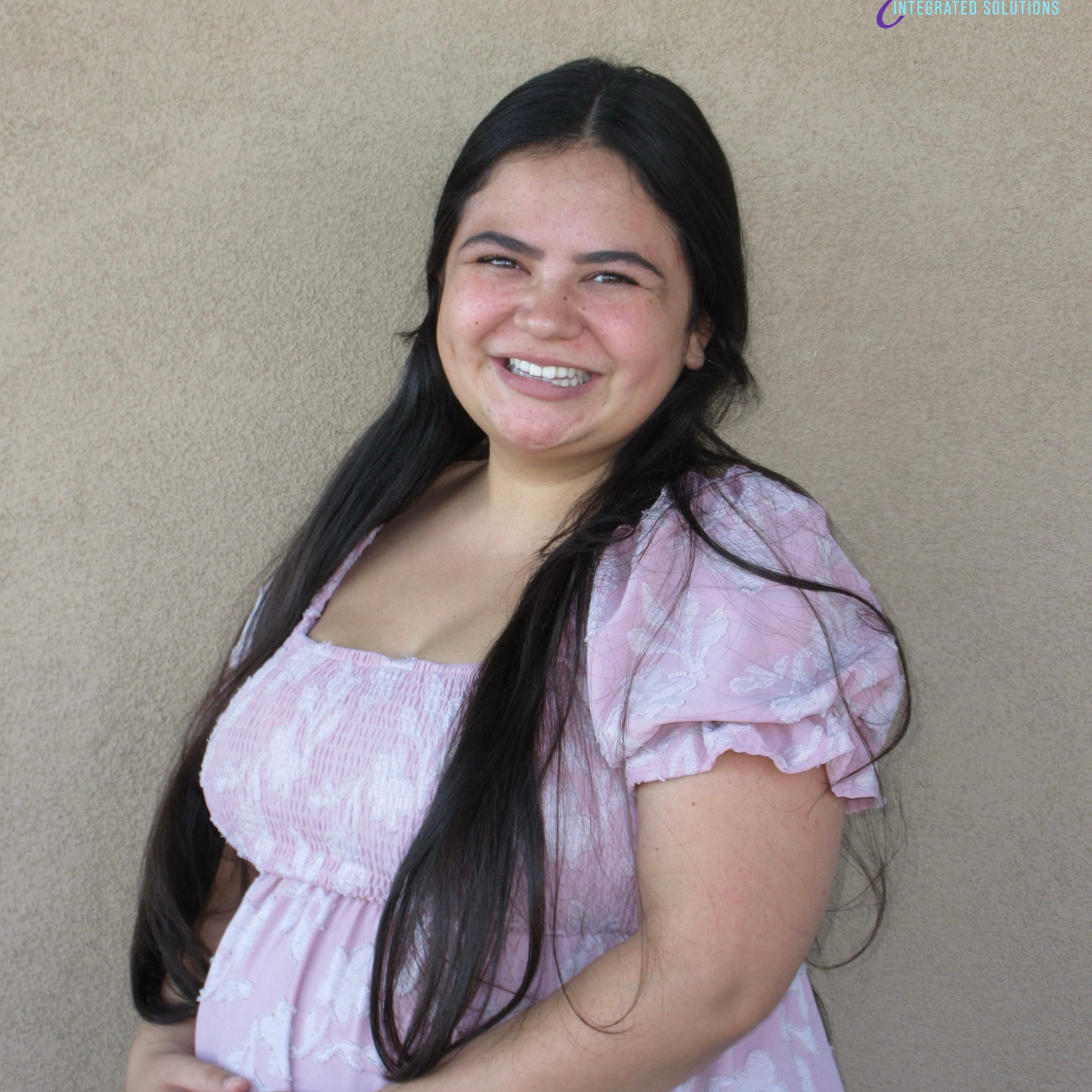 Smiling woman with long dark hair in a pink floral dress standing against beige wall with Grace logo in the top right corner.