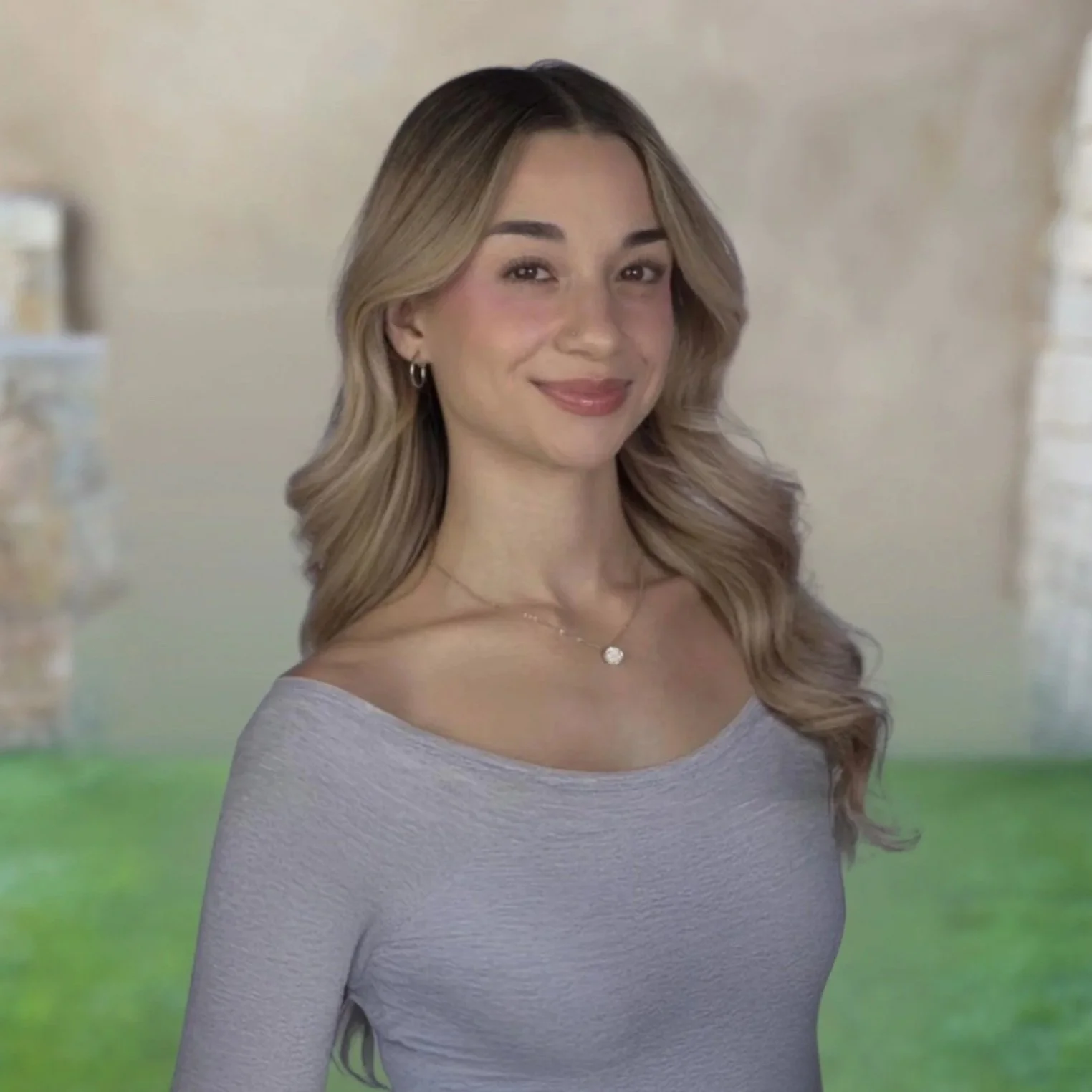 A woman with long, wavy blonde hair smiling, wearing a light gray, long-sleeved top, a necklace, and earrings, standing outdoors with a blurred background of grass, brick columns, and a beige wall.