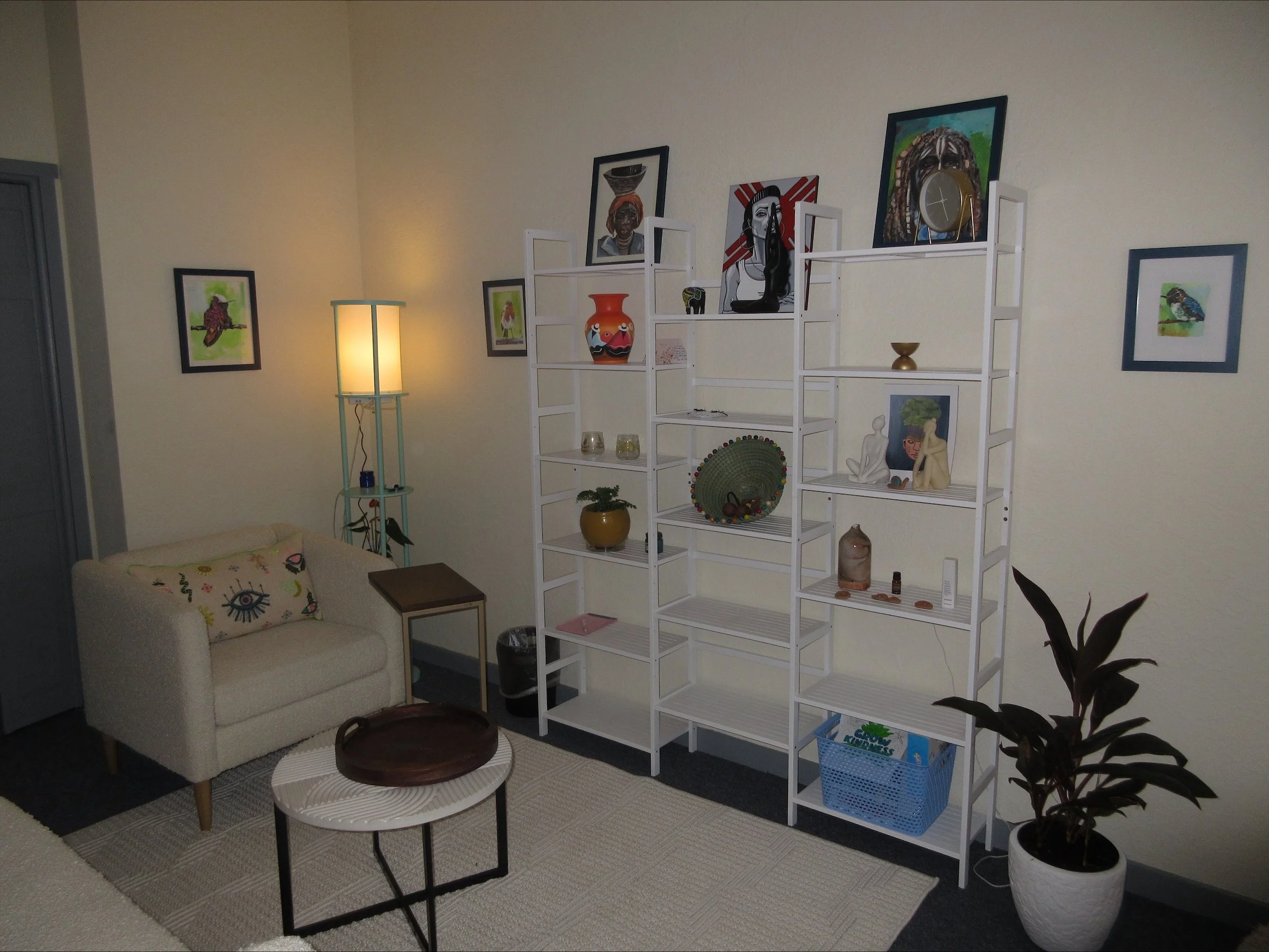 A cozy living room corner with a small beige couch, a round white coffee table with a wooden tray, a tall lit floor lamp, a white shelving unit with assorted art pieces, vases, and decorative objects, framed wall art, and a potted plant in a white po