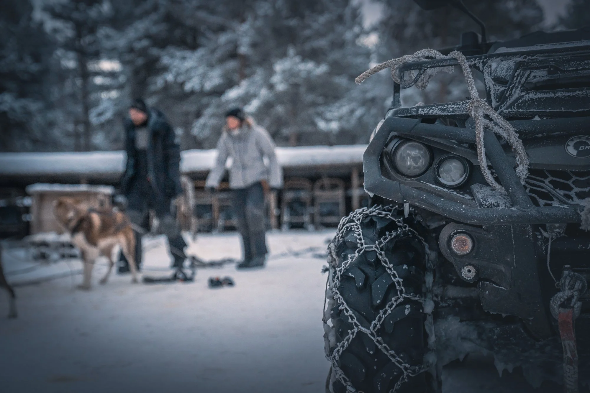Nærbilde av et snødekt terrengkjøretøy med kjettinger på dekkene, med to personer og en hund i bakgrunnen i et snødekt landskap om vinteren.