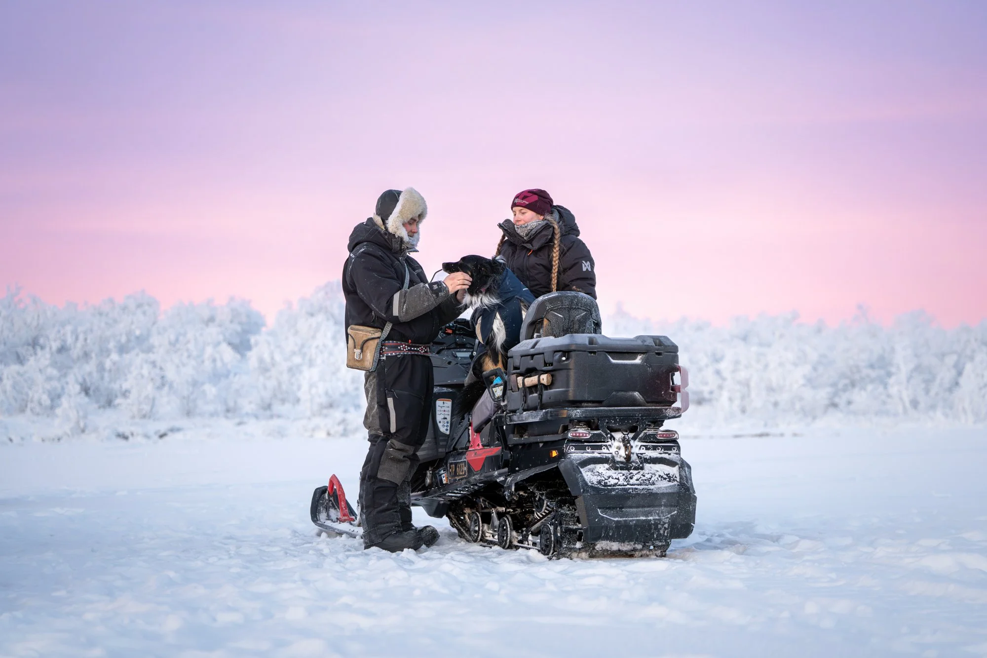 To personer i vinterutstyr står ved en snøscooter i et snødekt landskap, med en rosa og lilla himmel i bakgrunnen.
