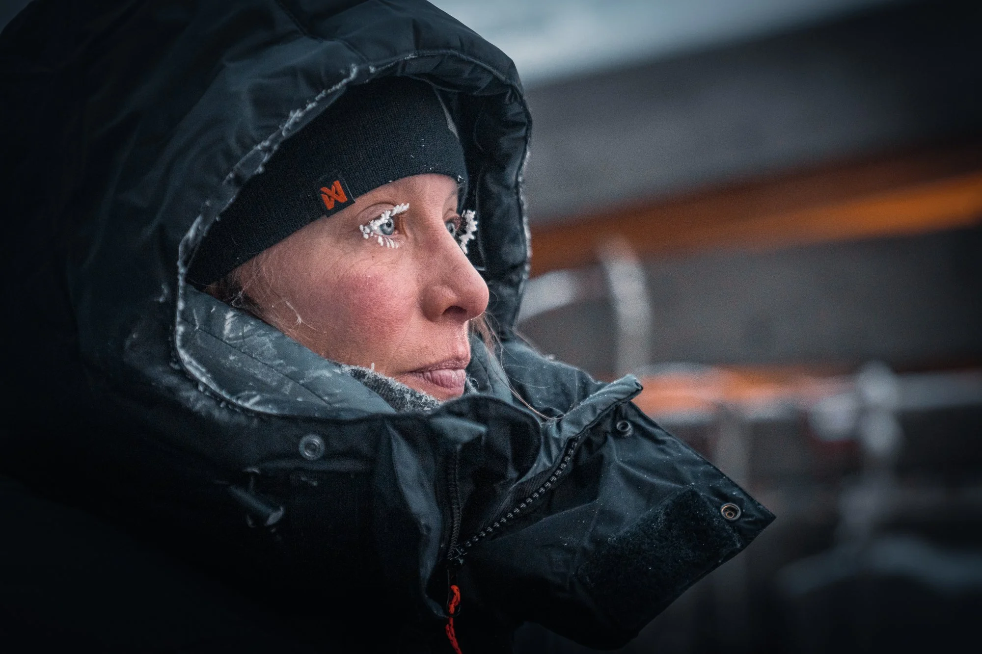 En kvinne i vintertøy, inkludert en svart lue med oransje logo og en svart hettejakke, ser til siden utendørs i kaldt vær med snø på øyevippene.