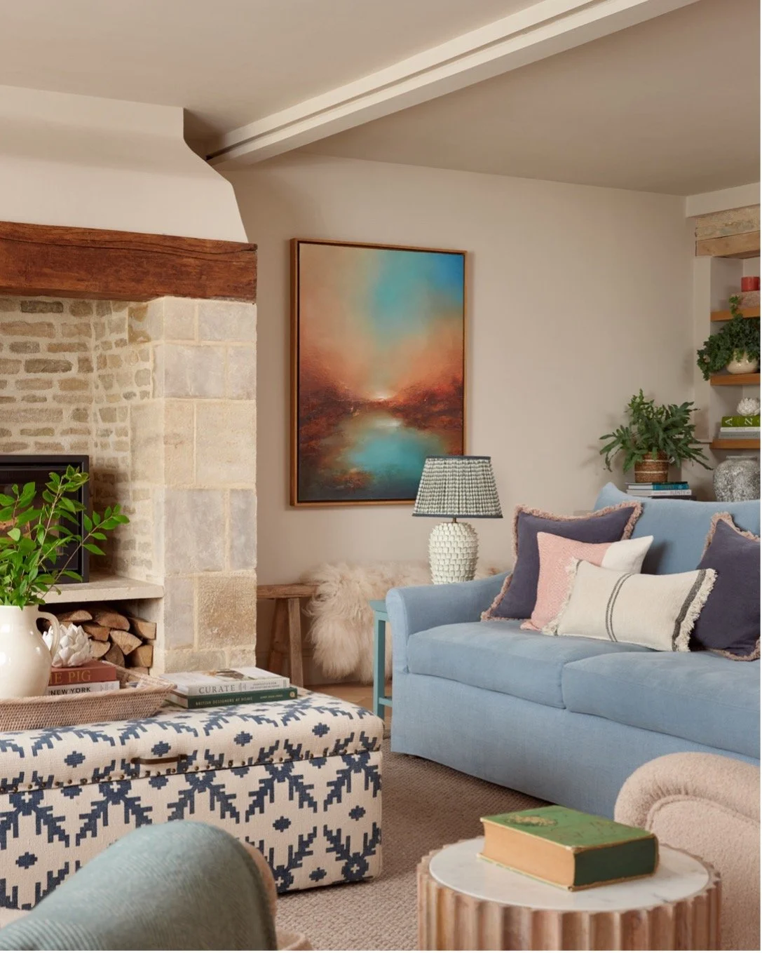 Cozy Cotswold sitting room with stone fireplace, log burner, blue sofa with coral and navy cushions, abstract landscape artwork by Jude McKenna, table lamp, ottoman and various plants.