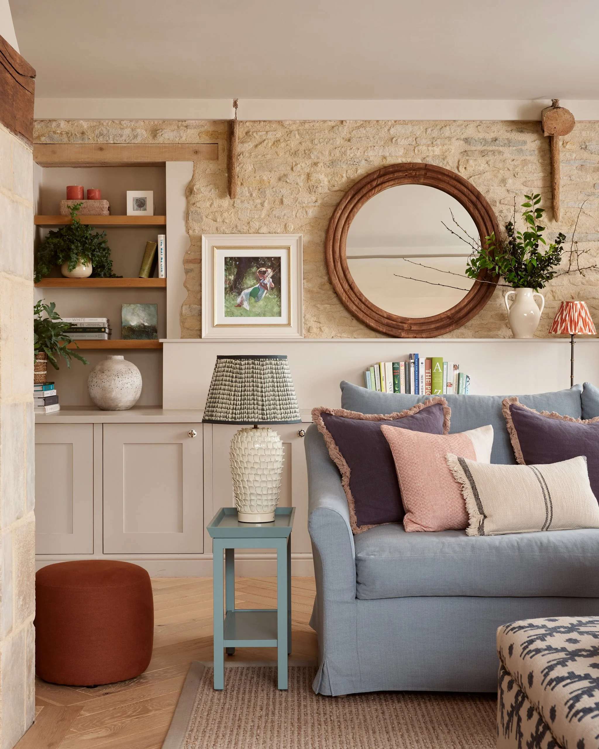 Beautifully styled sitting room in a Cotswold barn conversion. Blue sofa, assorted throw pillows, a Neptune side table with a textured Pooky lamp, a sideboard with decorative items and books, a large round mirror, and art.