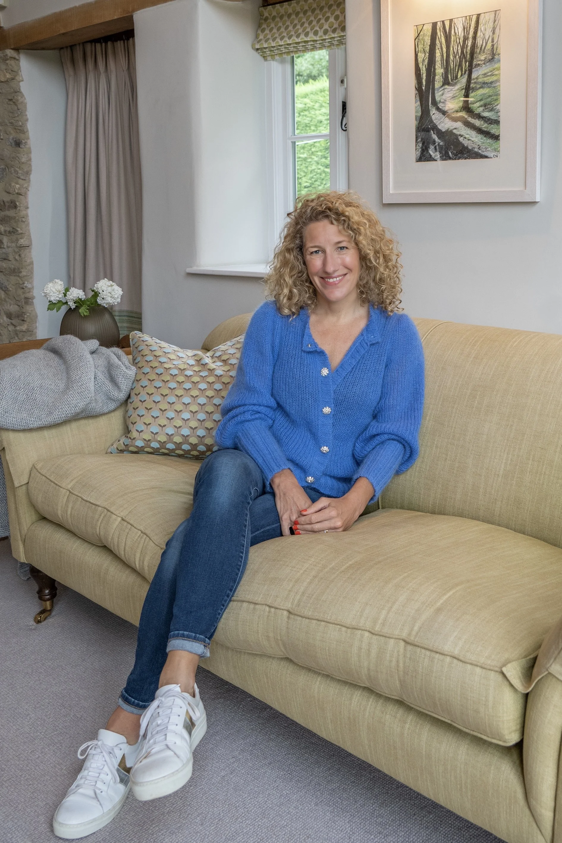 Woman sitting on beige sofa in a cozy living room, wearing a blue cardigan and jeans, with a window and wall art in the background.