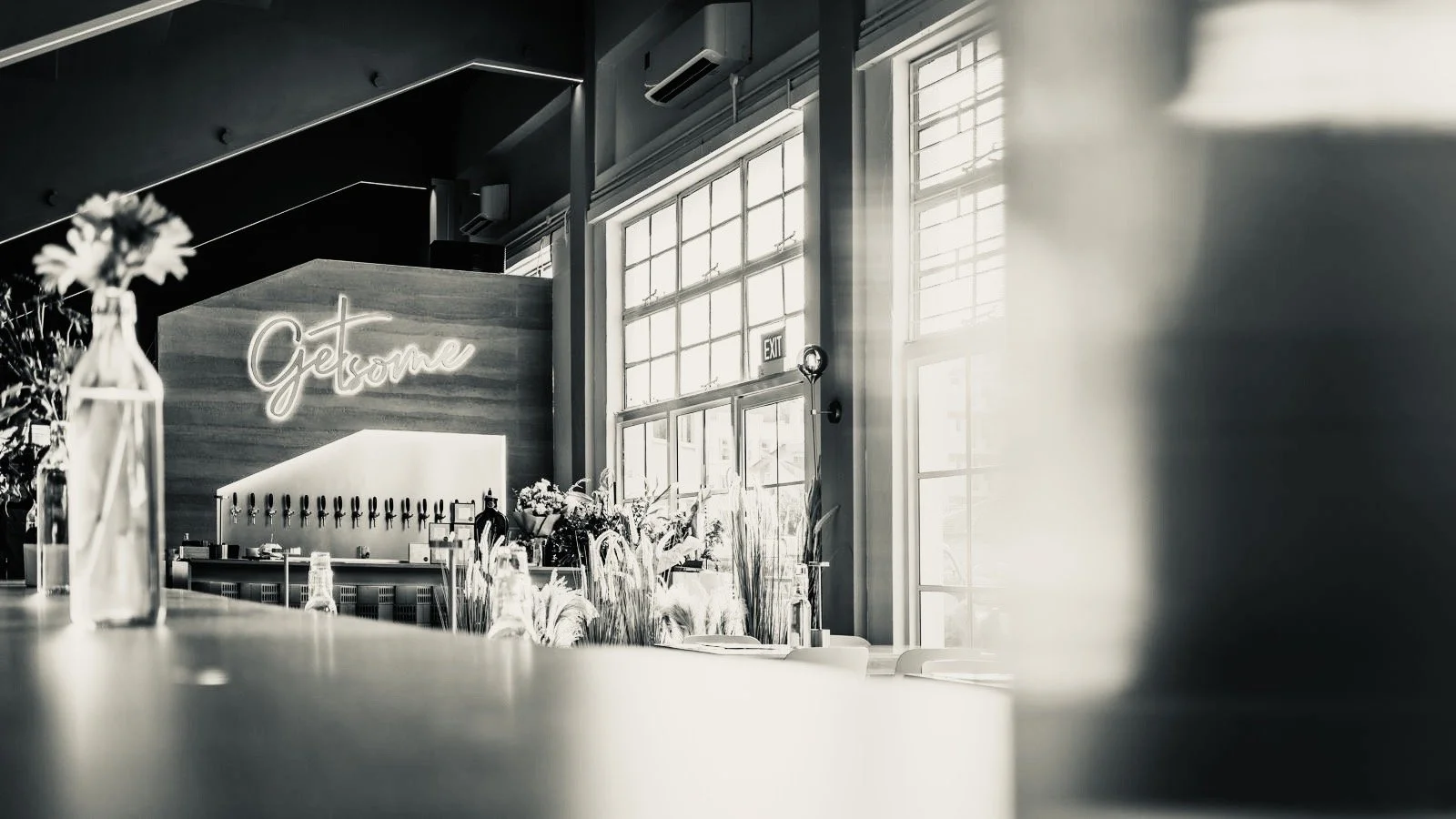 Interior of a modern café or bar with large windows, a neon sign that says 'Getome', and a counter with drinks taps and flowers.