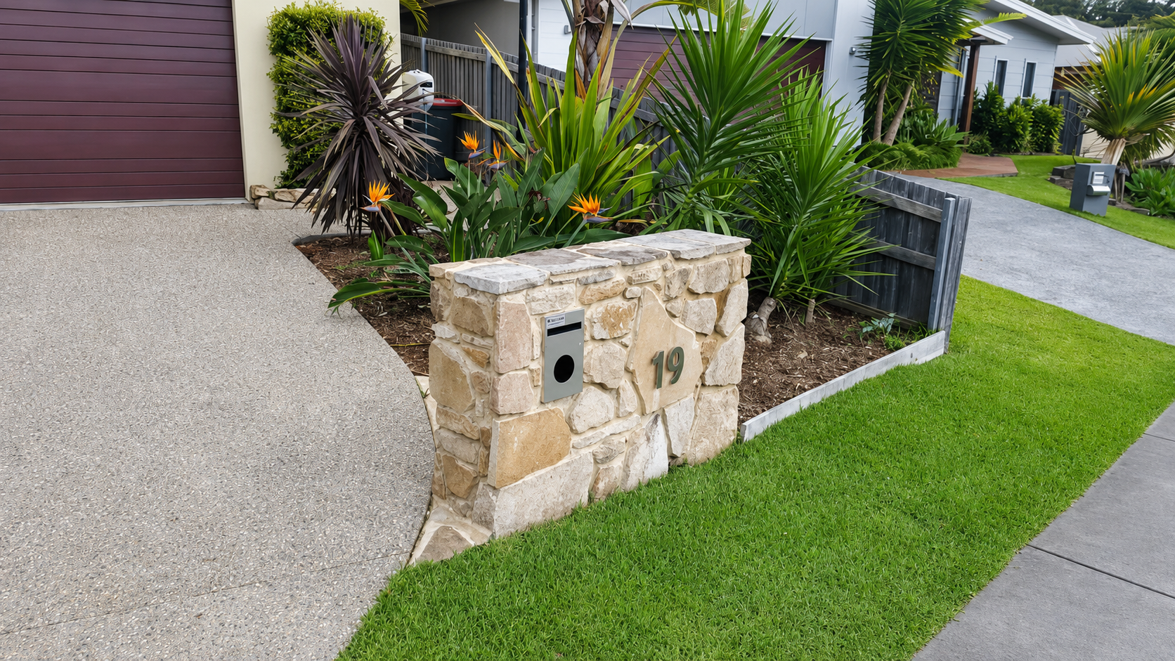 A stone mailbox with house number 19, surrounded by lush green plants and a well-maintained lawn in a residential neighborhood.