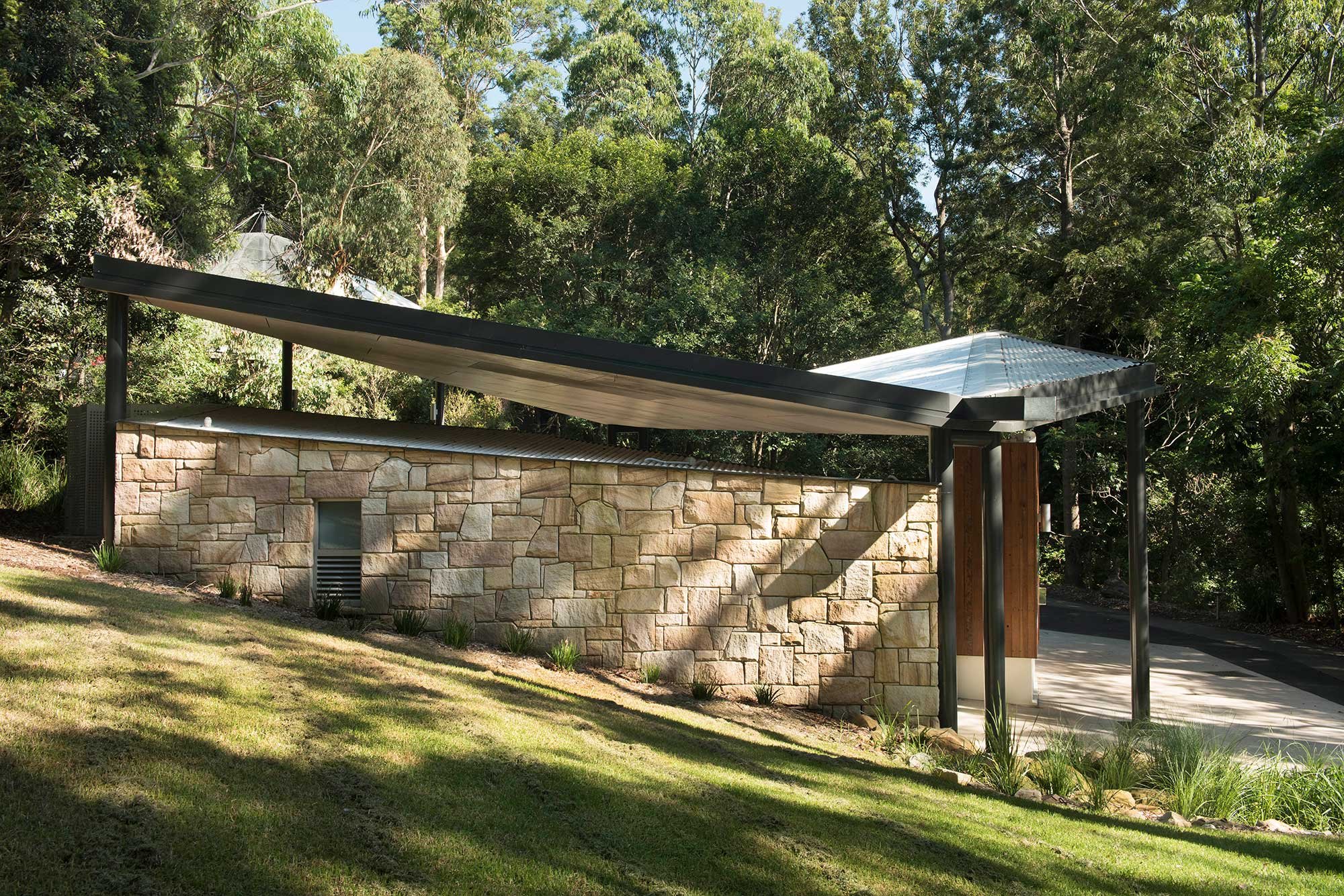 Exterior of a modern building with a stone wall, metal roof, and surrounding trees and grass.