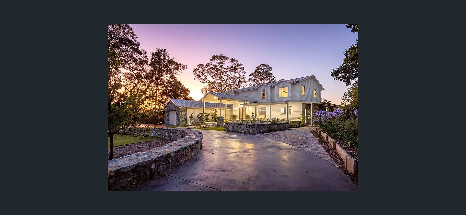 A modern two-story house with a spacious driveway, surrounded by trees and flowering plants at sunset.