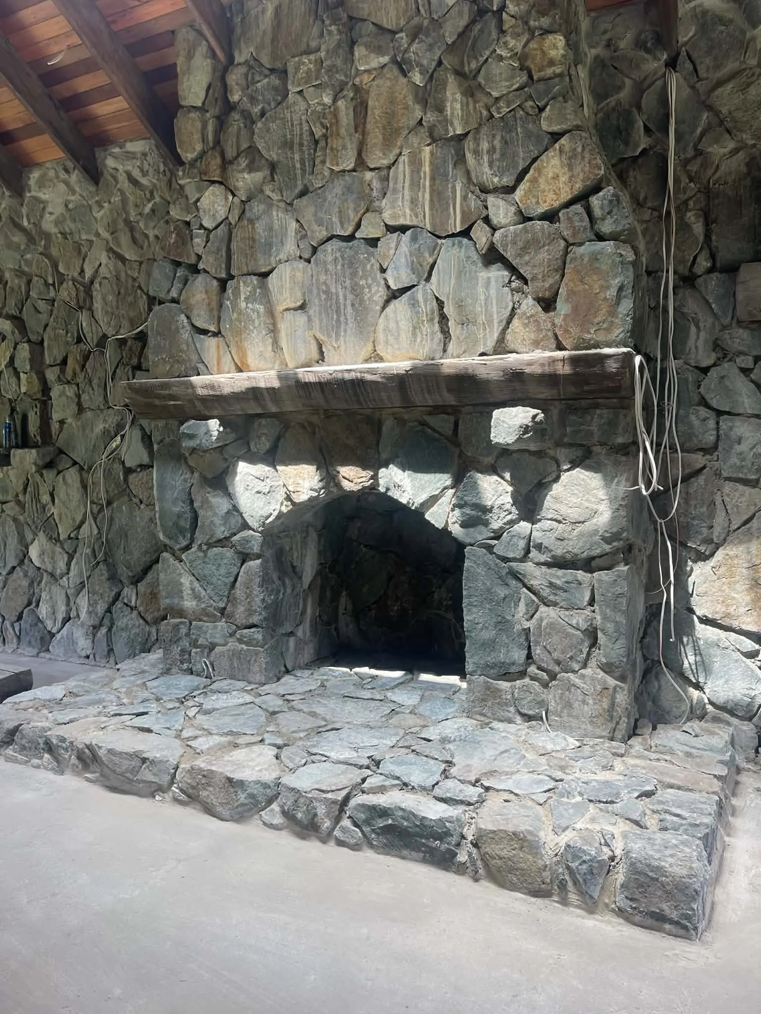A stone fireplace with a wooden mantel, surrounded by large irregular stones, in a rustic setting.