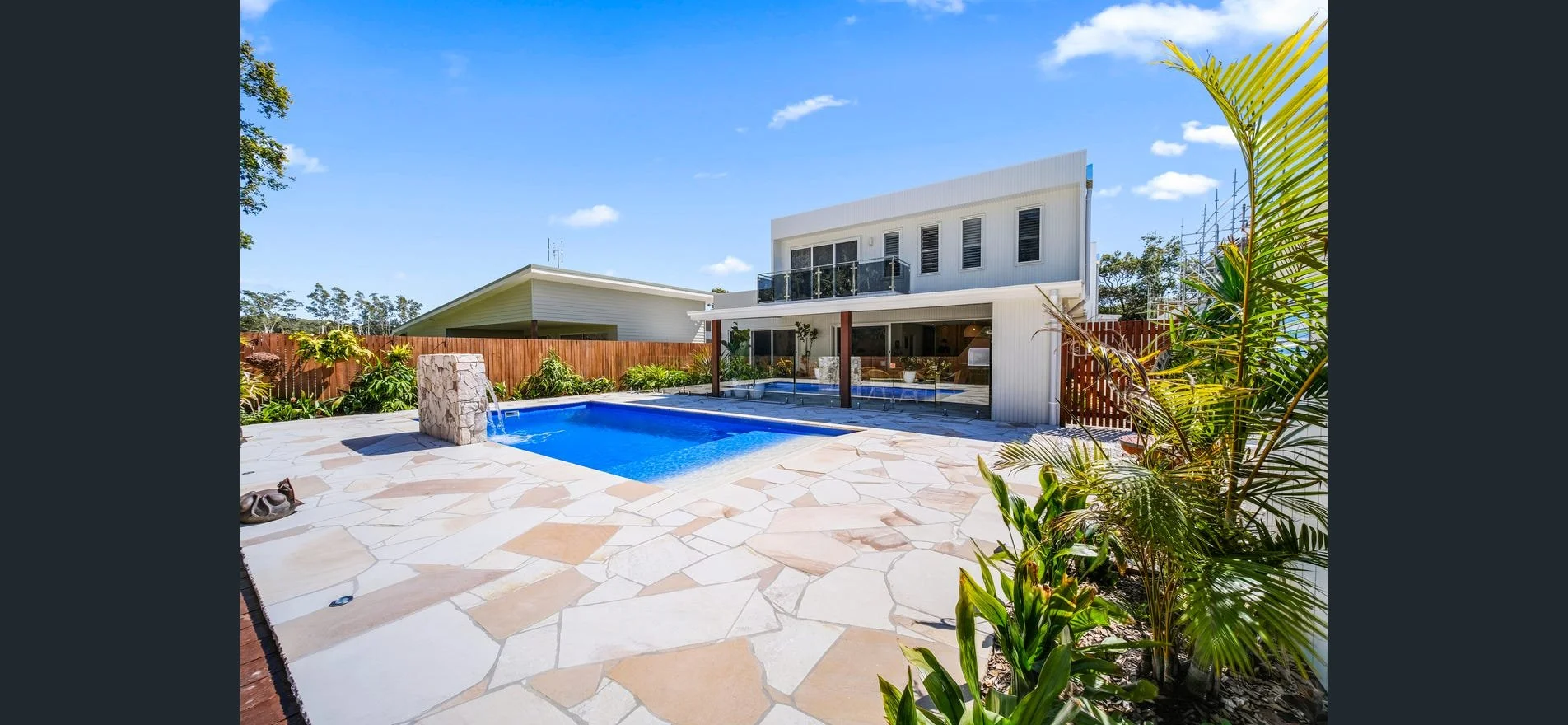 Modern two-story house with a backyard swimming pool, surrounded by a stone patio and lush green plants, under a bright blue sky.