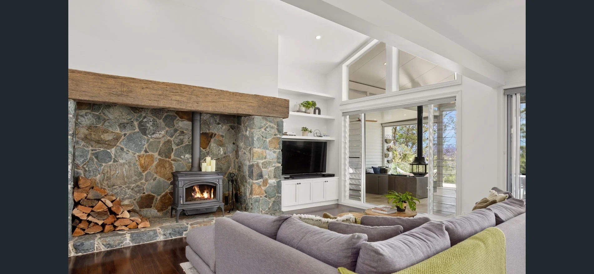 Modern living room with a stone and wood fireplace, a gray sofa with a yellow cushion, a white TV unit, and a view into an outdoor screened porch with trees outside.