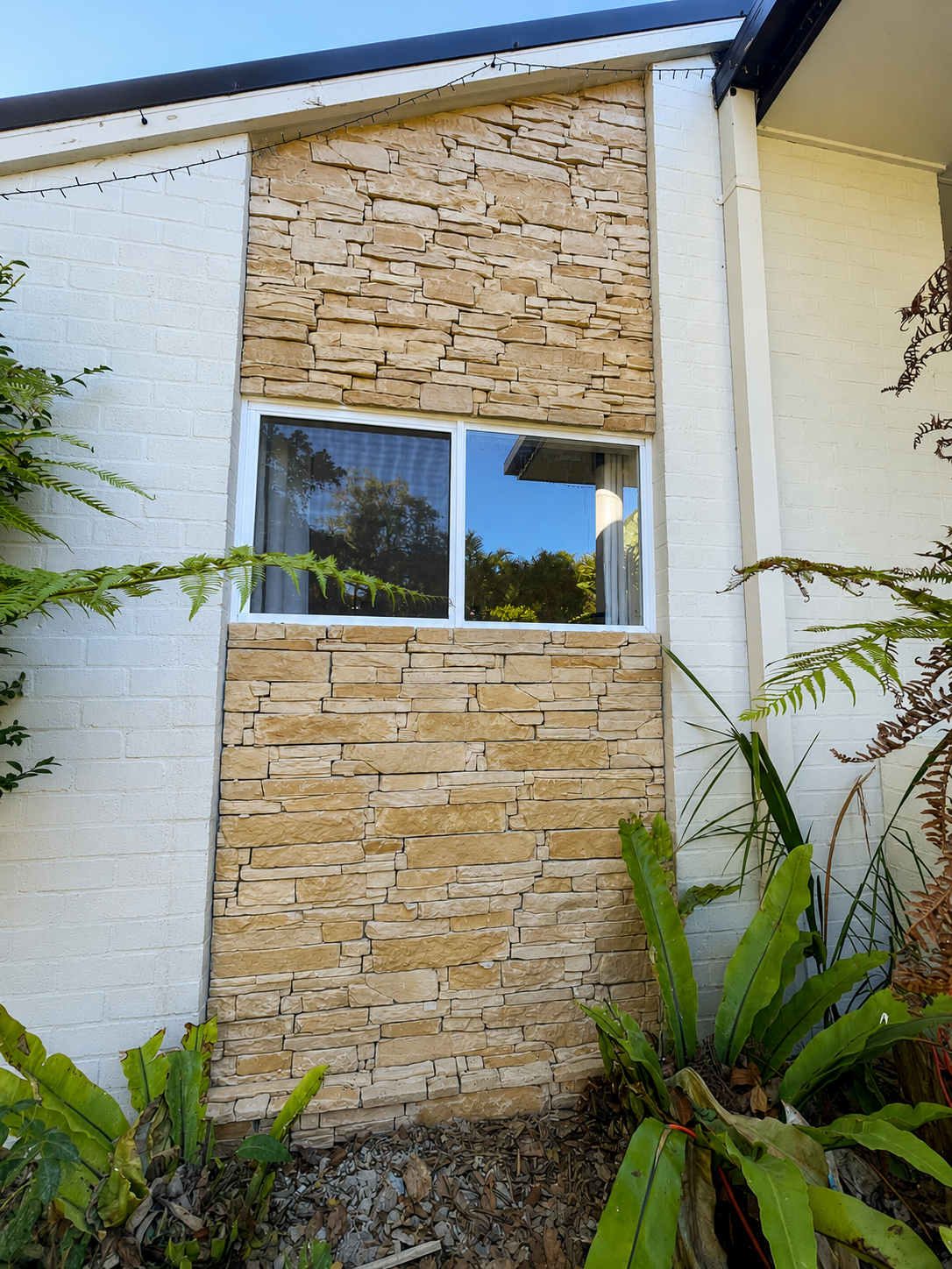 Close-up of a house exterior featuring white brick walls, beige stone accents around a window, and green plants in the foreground.