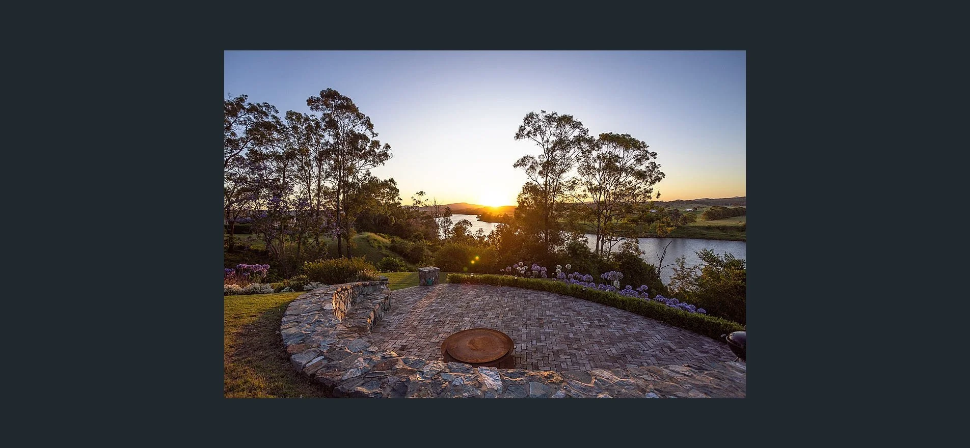 Sunset over a river viewed from a stone patio with a curved stone bench and flowering bushes.