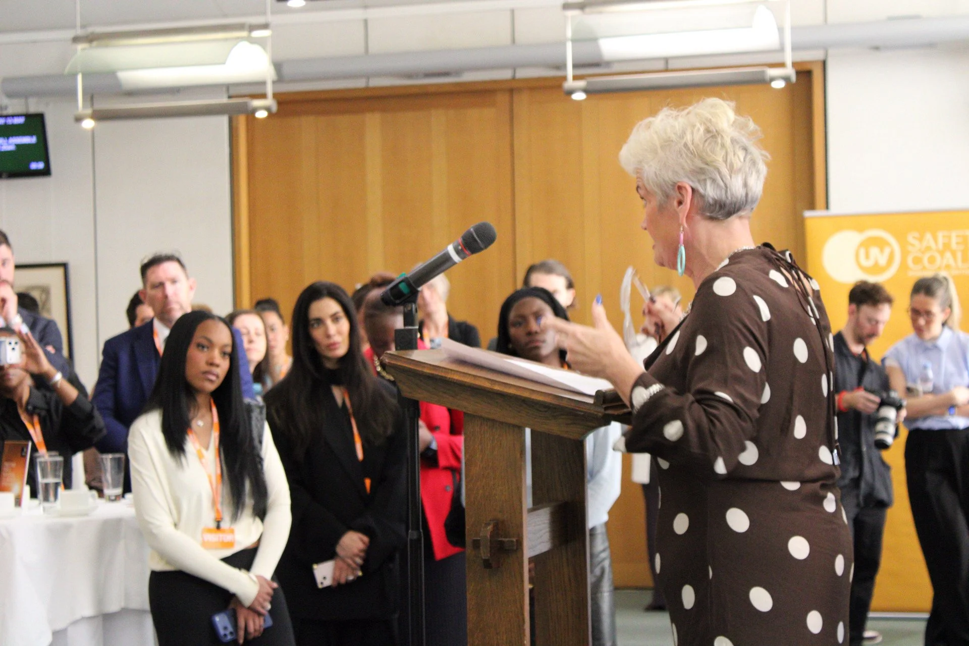 A woman with short gray hair and turquoise earrings, wearing a brown polka dot top, is standing at a podium speaking to an audience at a conference. The audience consists of diverse people, some with cameras, listening attentively. There is a sign in