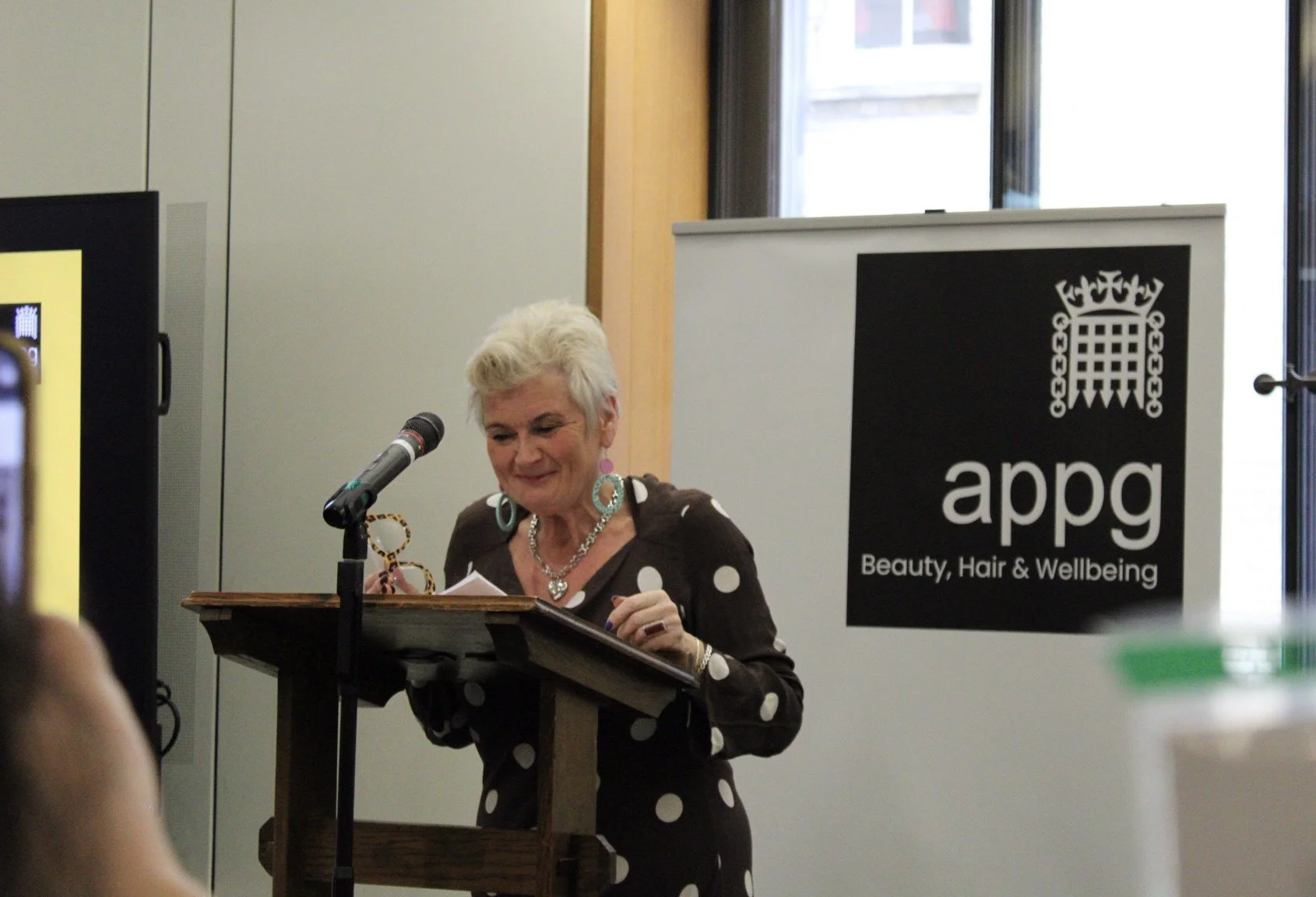 A woman with short white hair, wearing a dark polka dot dress and jewelry, stands behind a wooden podium speaking into a microphone at an event. There is a black banner with white text and a logo to her right that reads 'apdg, Beauty, Hair & Wellbein