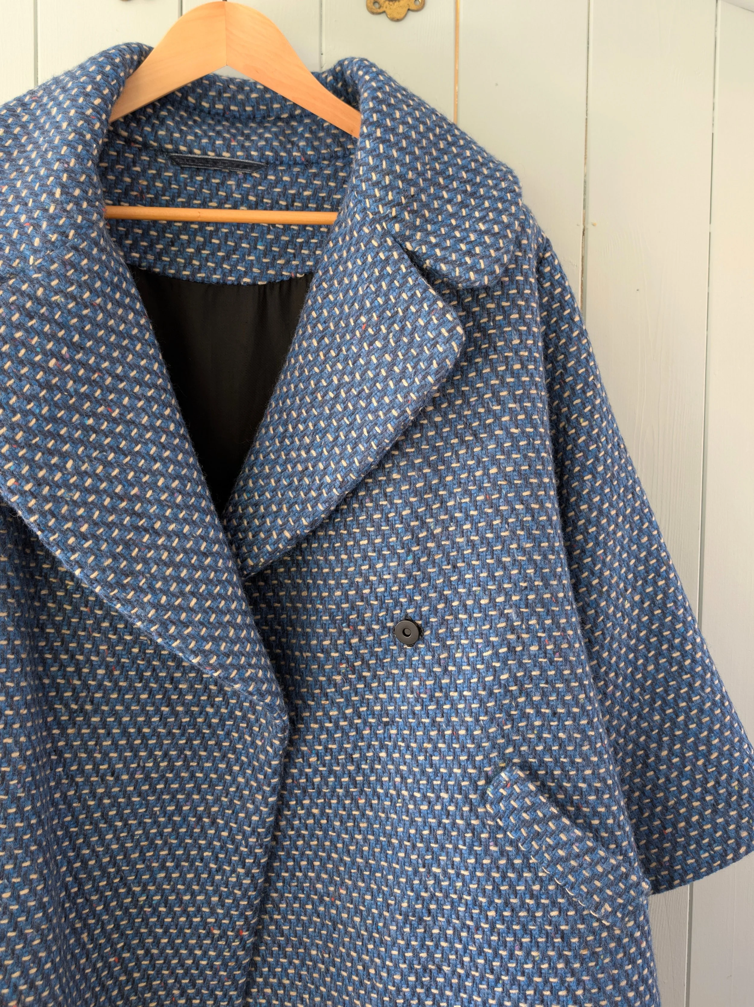Blue textured coat hanging on a wooden hanger against a light-colored wall.