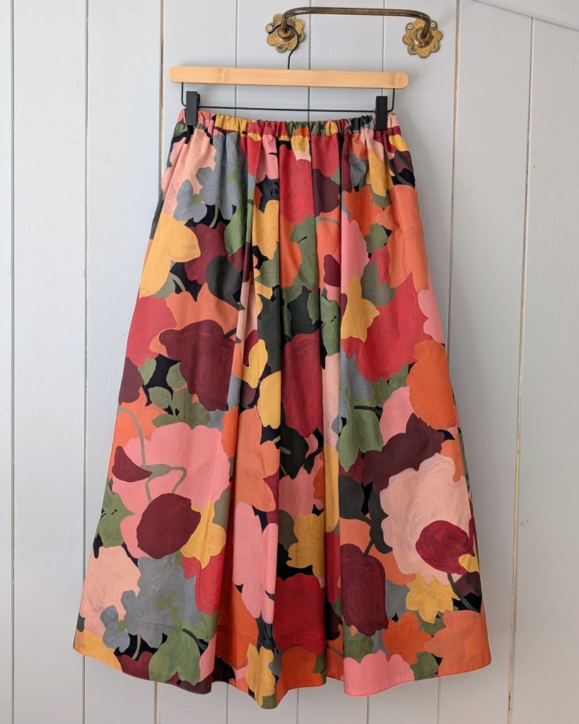 A colorful, floral-patterned maxi skirt hanging on a wooden hanger against a light gray wooden wall.