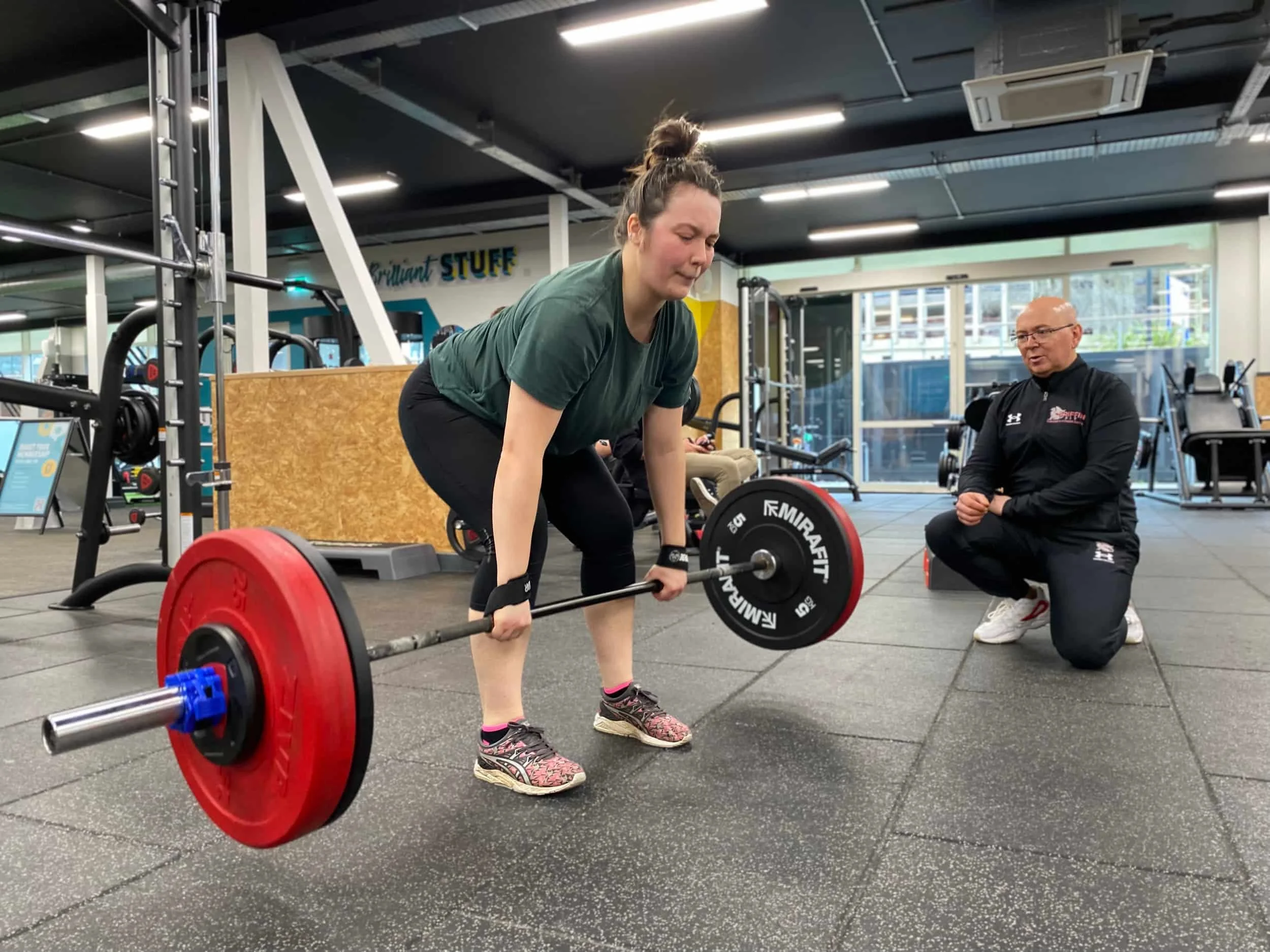 A woman lifts a barbell in a gym as her personal training Oxford coach kneels nearby, observing her form.